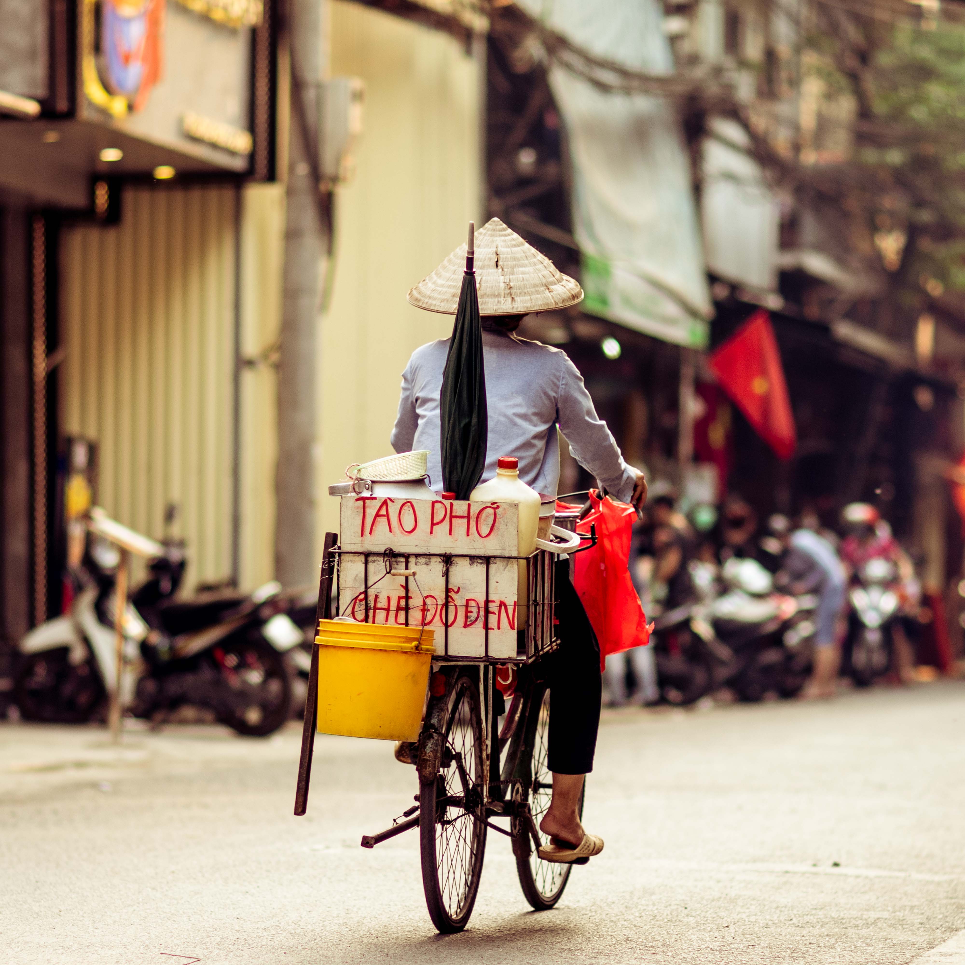 Vietnamese street vendor in a conical hat rides a bicycle loaded with 'Tào Phớ' and 'Chè Đỗ Đen' dessert coolers through a narrow Hanoi street.