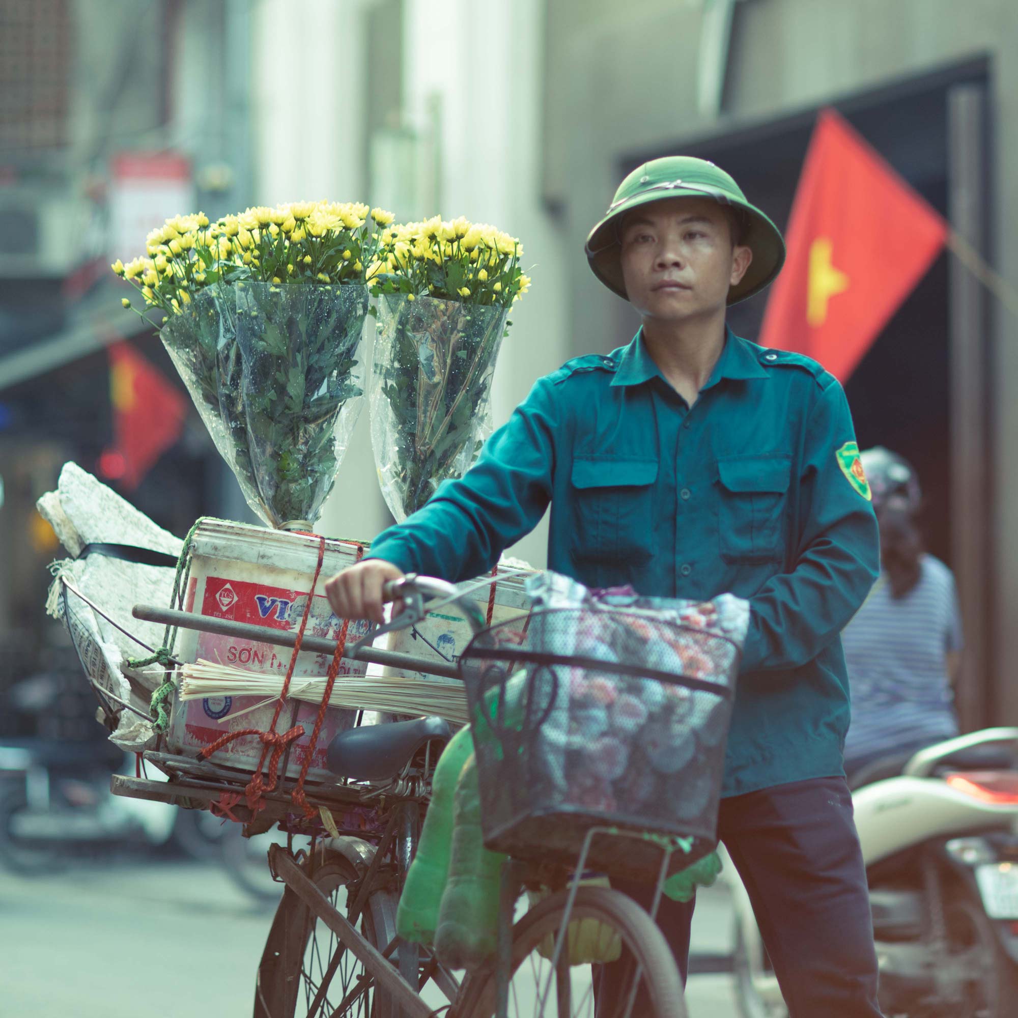 Street portrait of a Vietnamese man in a green pith helmet and uniform pushing a bicycle loaded with yellow chrysanthemum bouquets through a Hanoi street.