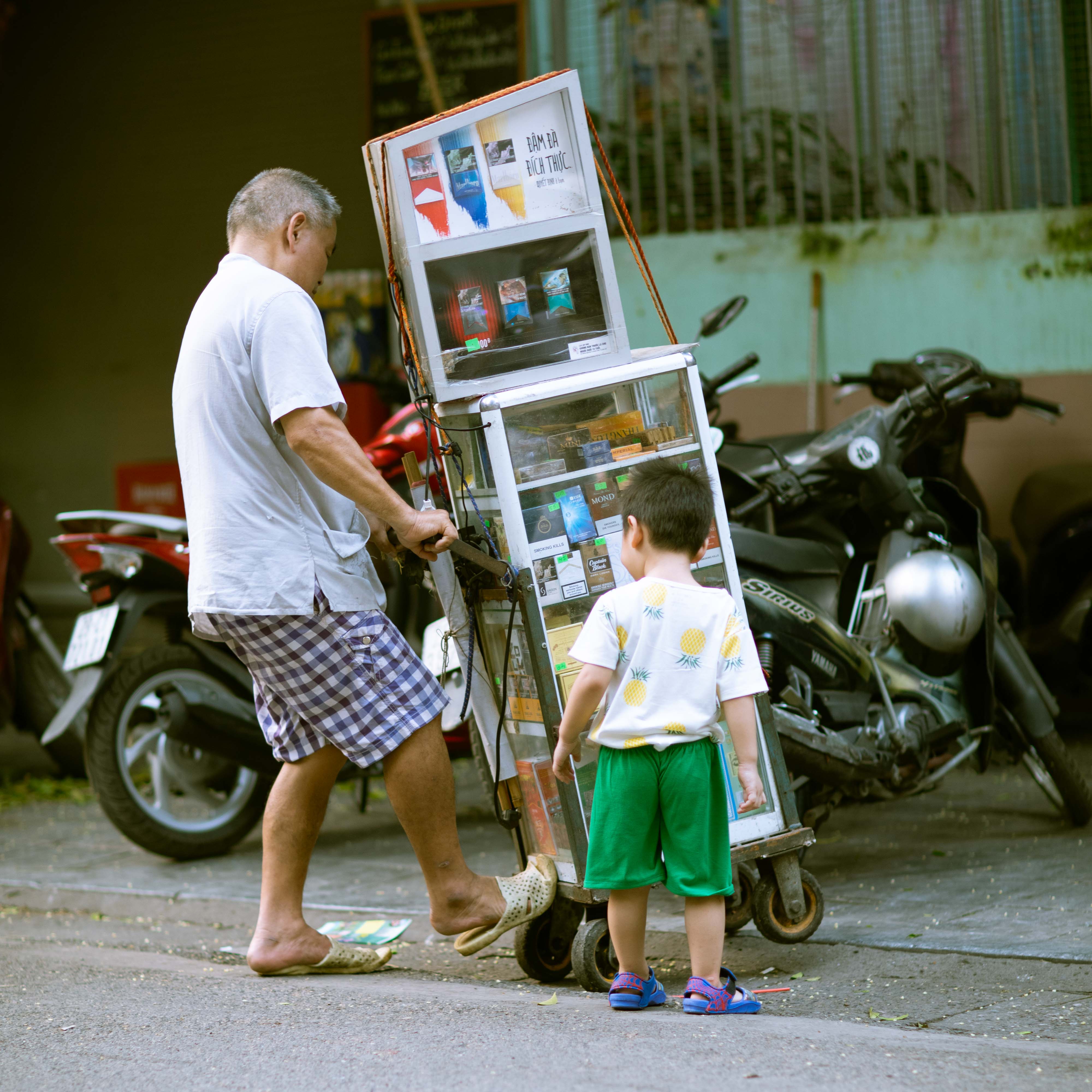 A Vietnamese shopkeeper wheels a mobile convenience cart along a motorbike-lined Hanoi sidewalk as a small boy in green shorts watches.