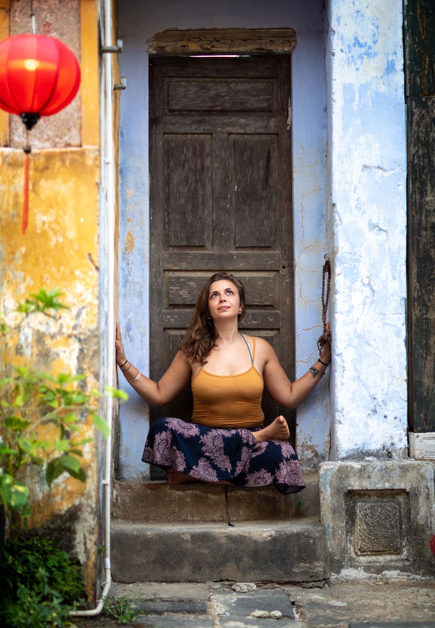 Emilia Semrau sits cross-legged in meditation inside a weathered blue and yellow doorway beside a red paper lantern in Hoi An, Vietnam.