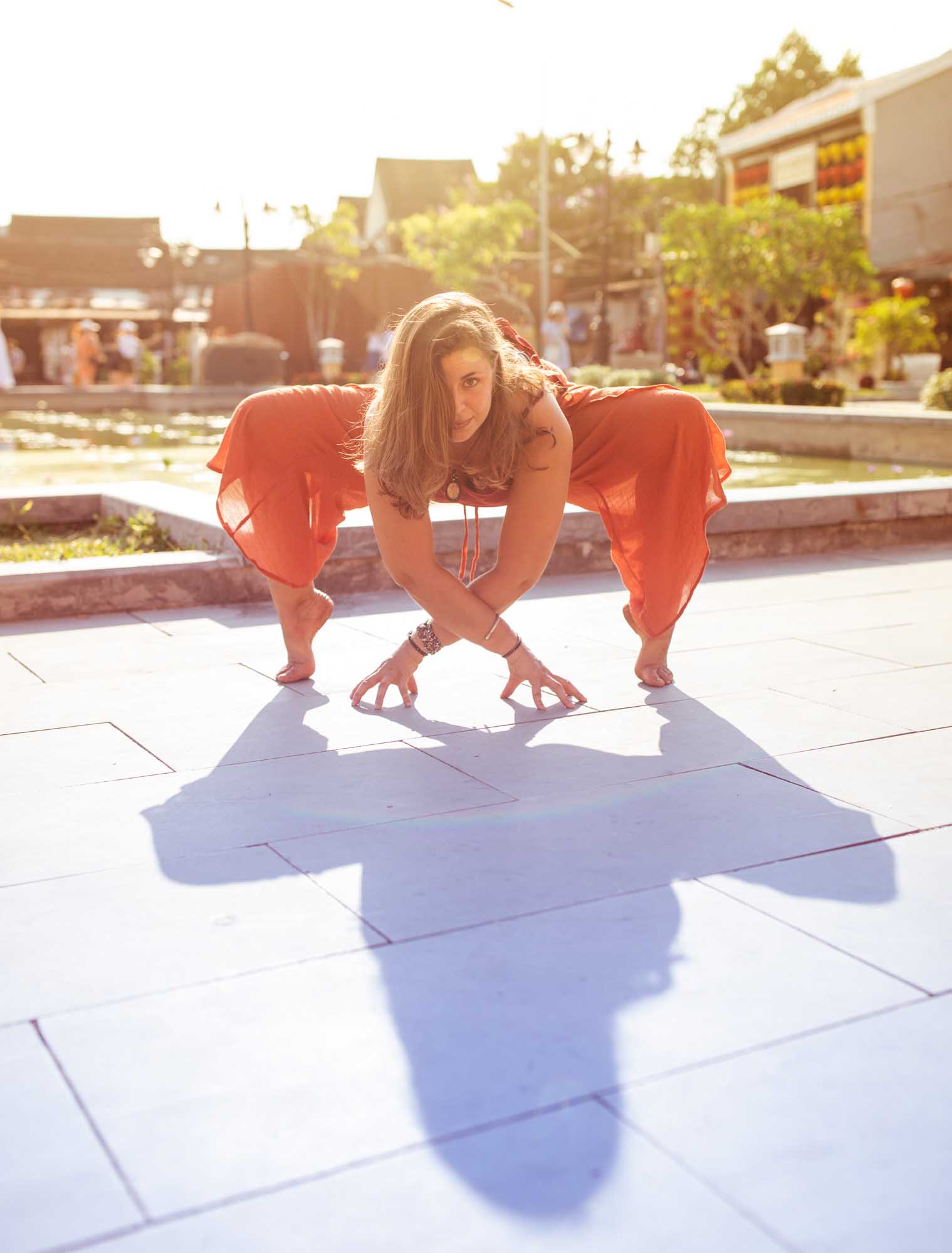 Emilia Semrau in a wide yogi squat on a sunlit temple plaza in Hoi An, Vietnam, wearing flowing orange pants.