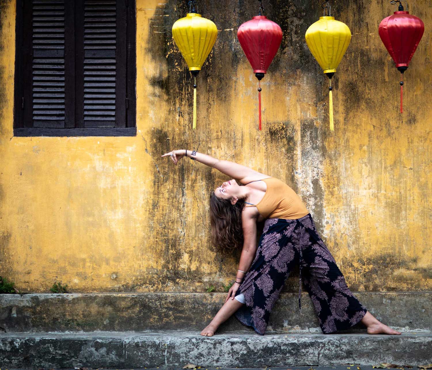 Emilia Semrau in a standing side-bend yoga pose against a yellow wall hung with red and yellow paper lanterns, Hoi An, Vietnam.