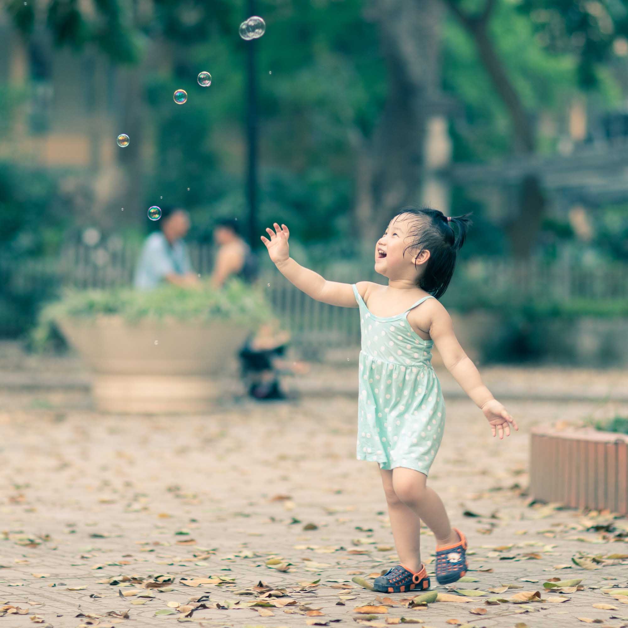 Street portrait of a toddler in a pale-green dress laughing and reaching for soap bubbles in a Hanoi park, Vietnam.