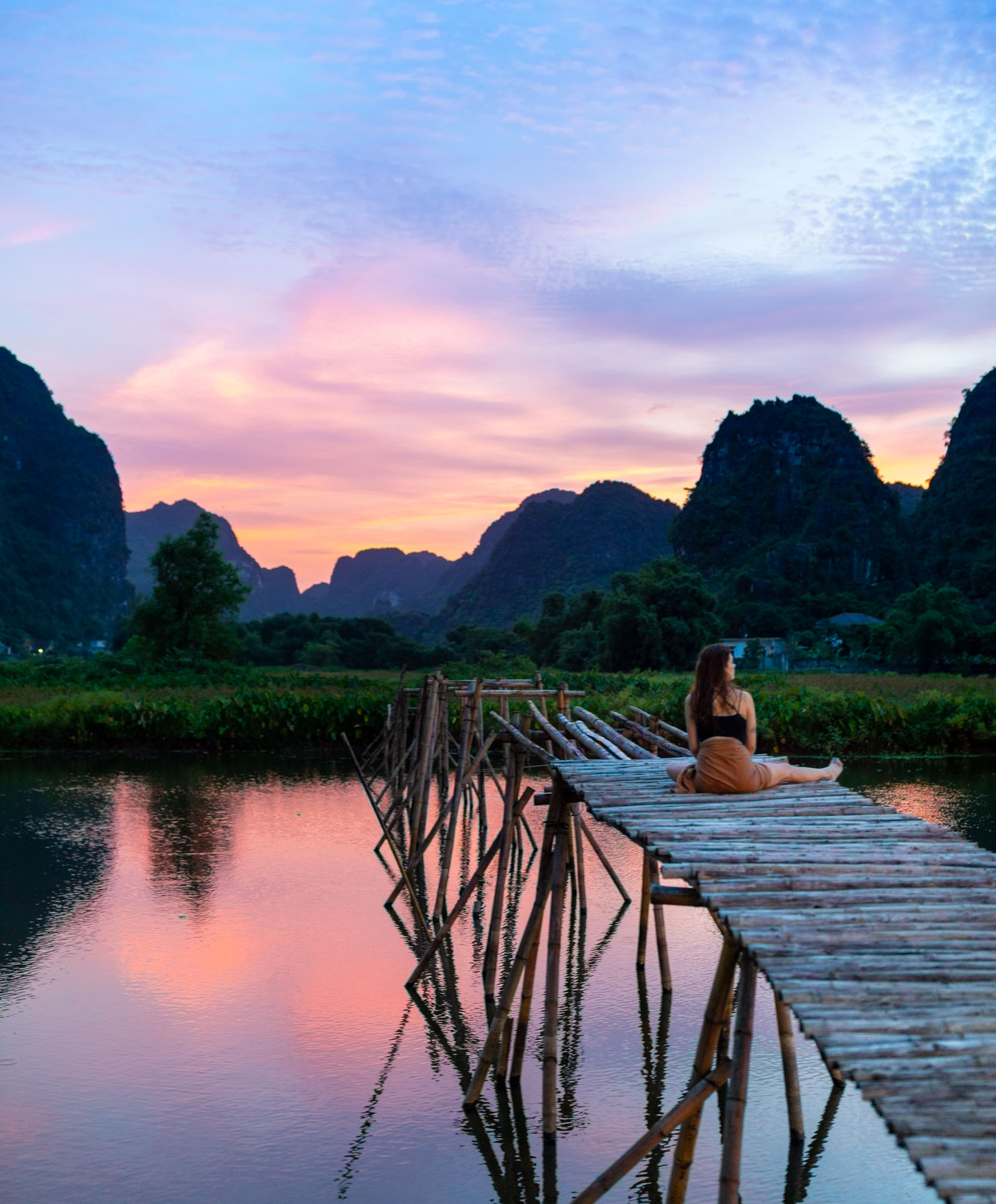 A woman sits on a long bamboo walkway across still water, watching a pink-and-purple sunset over the limestone karsts of Trang An, Vietnam.
