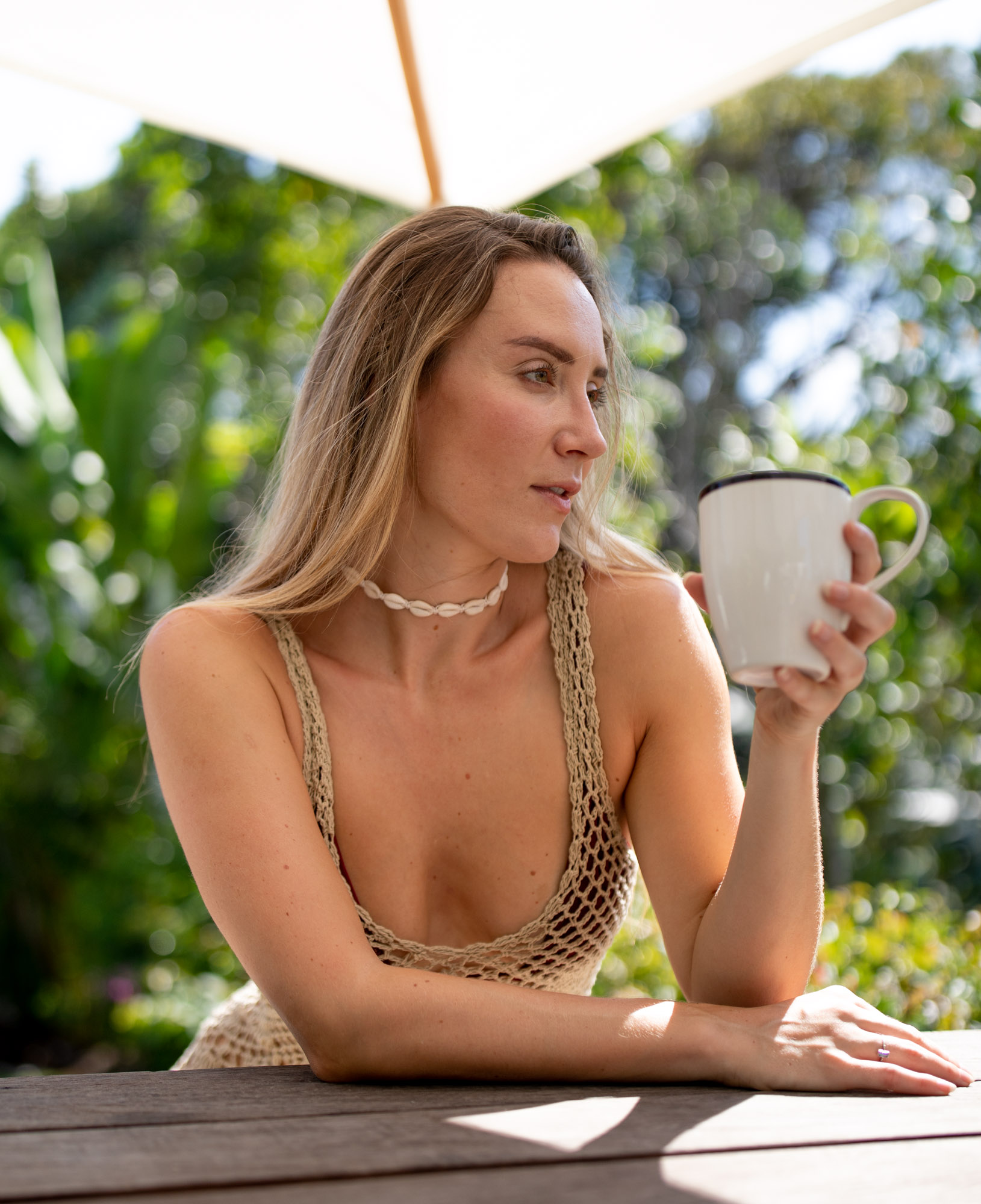 Blonde woman in a crochet tank holds a coffee mug at an outdoor wooden table under a sun umbrella in a Canggu, Bali garden.