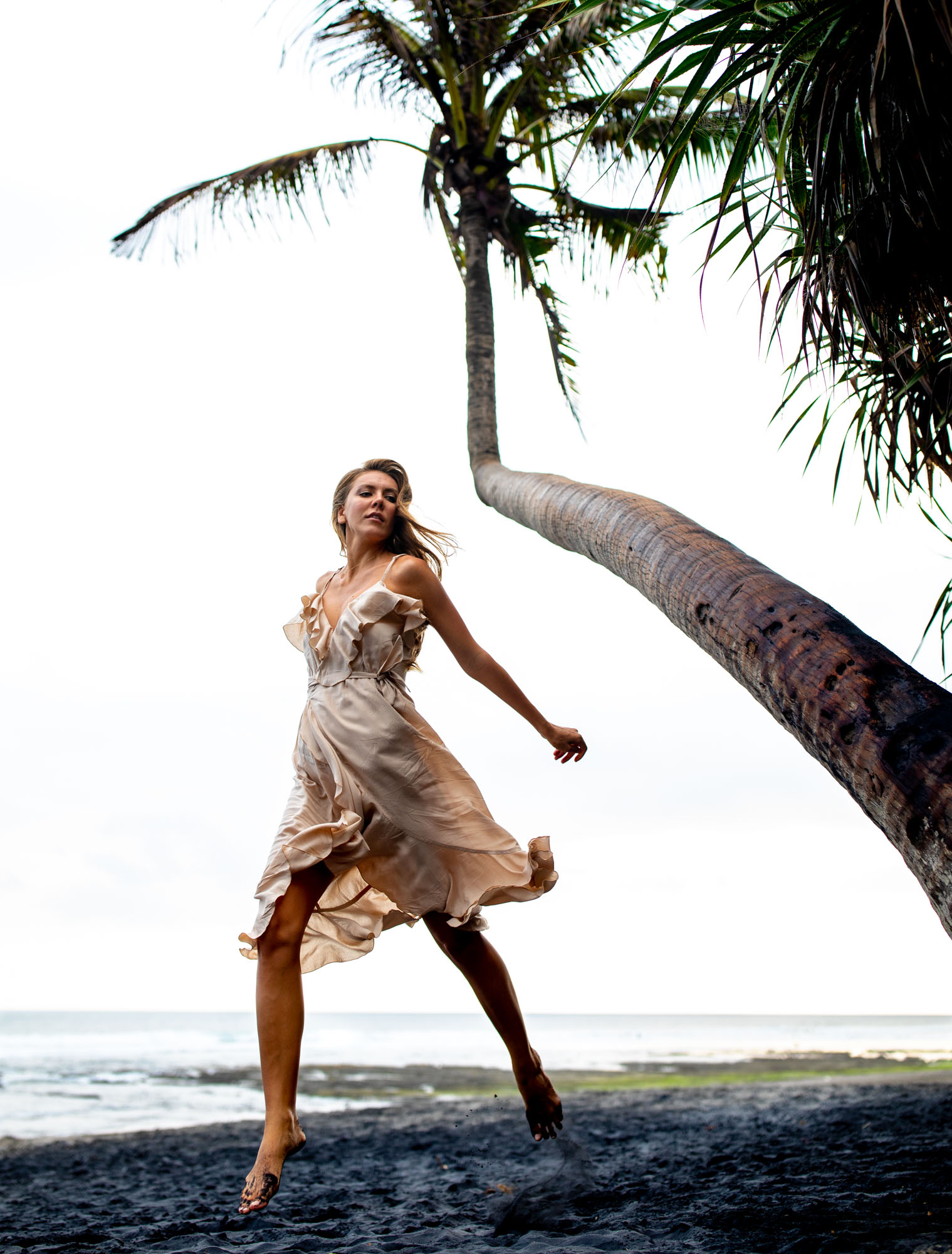 Model @alexa_air mid-jump on a black-sand beach under a leaning coconut palm, wearing a flowing nude silk dress — Canggu, Bali.
