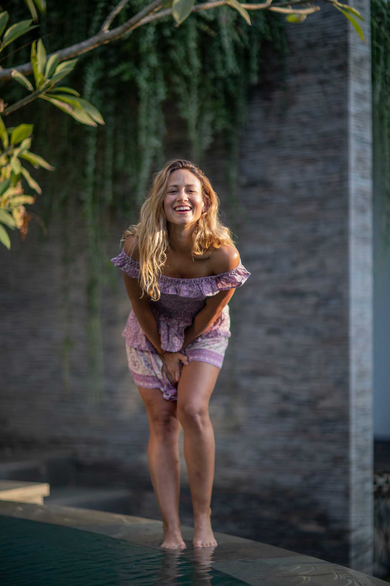 Blonde woman in a lilac off-shoulder dress laughs barefoot at the pool edge under hanging vines — Toraja Villa, Canggu, Bali.
