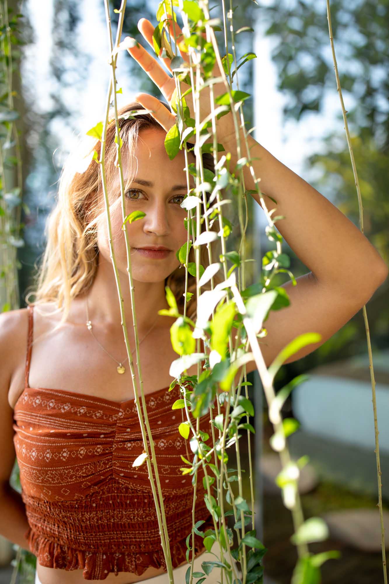 Portrait of a blonde woman in a rust-printed smocked crop top peering through a curtain of hanging vines — Toraja Villa, Canggu, Bali.