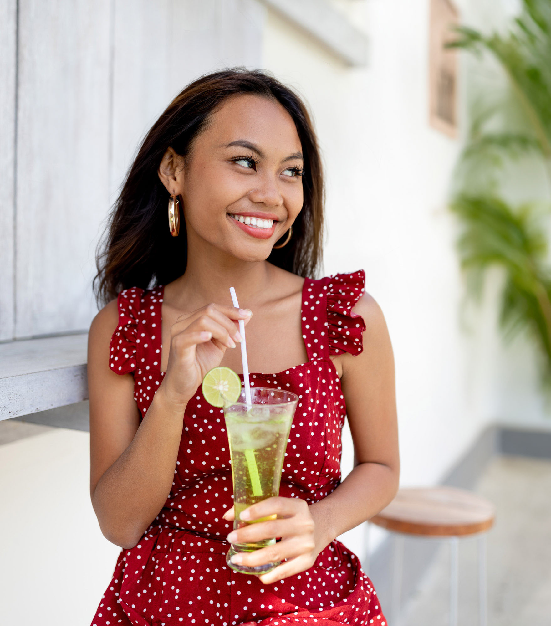 Hailey Ori laughs on a villa patio in Jimbaran, Bali, sipping a cocktail through a straw, wearing a red polka-dot ruffle dress.