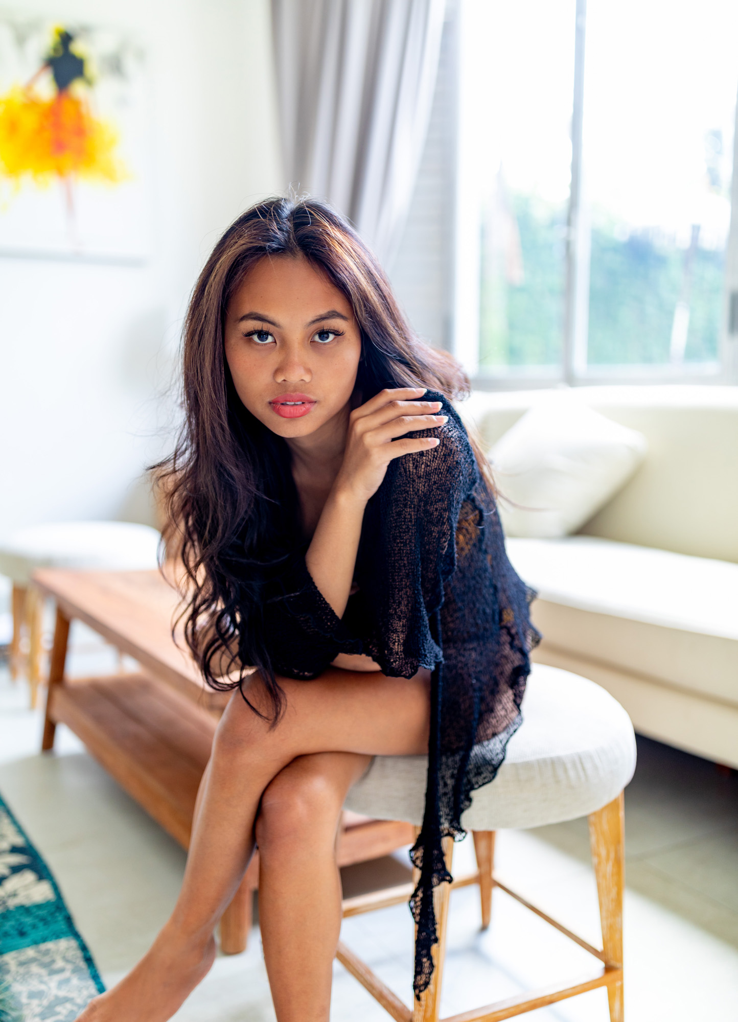 Hailey Ori sits on a stool in a sunlit villa living room in Bali, wearing a sheer black knit wrap.