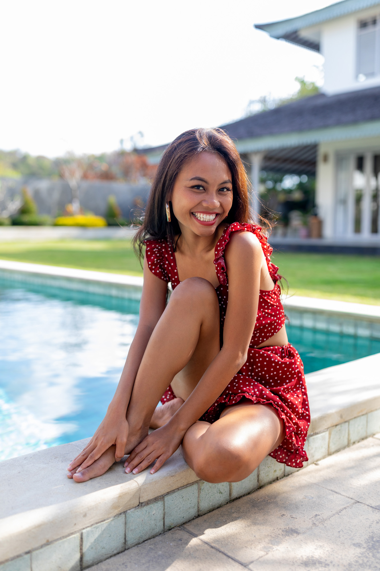 Hailey Ori sits on the edge of a villa pool in Jimbaran, Bali, laughing toward the camera in a red polka-dot dress.