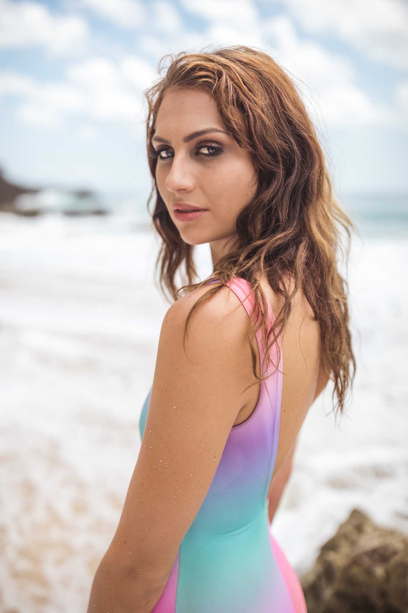 A redhead in a pink-and-teal ombré one-piece swimsuit looks over her shoulder with crashing surf behind her — Bali, Indonesia.