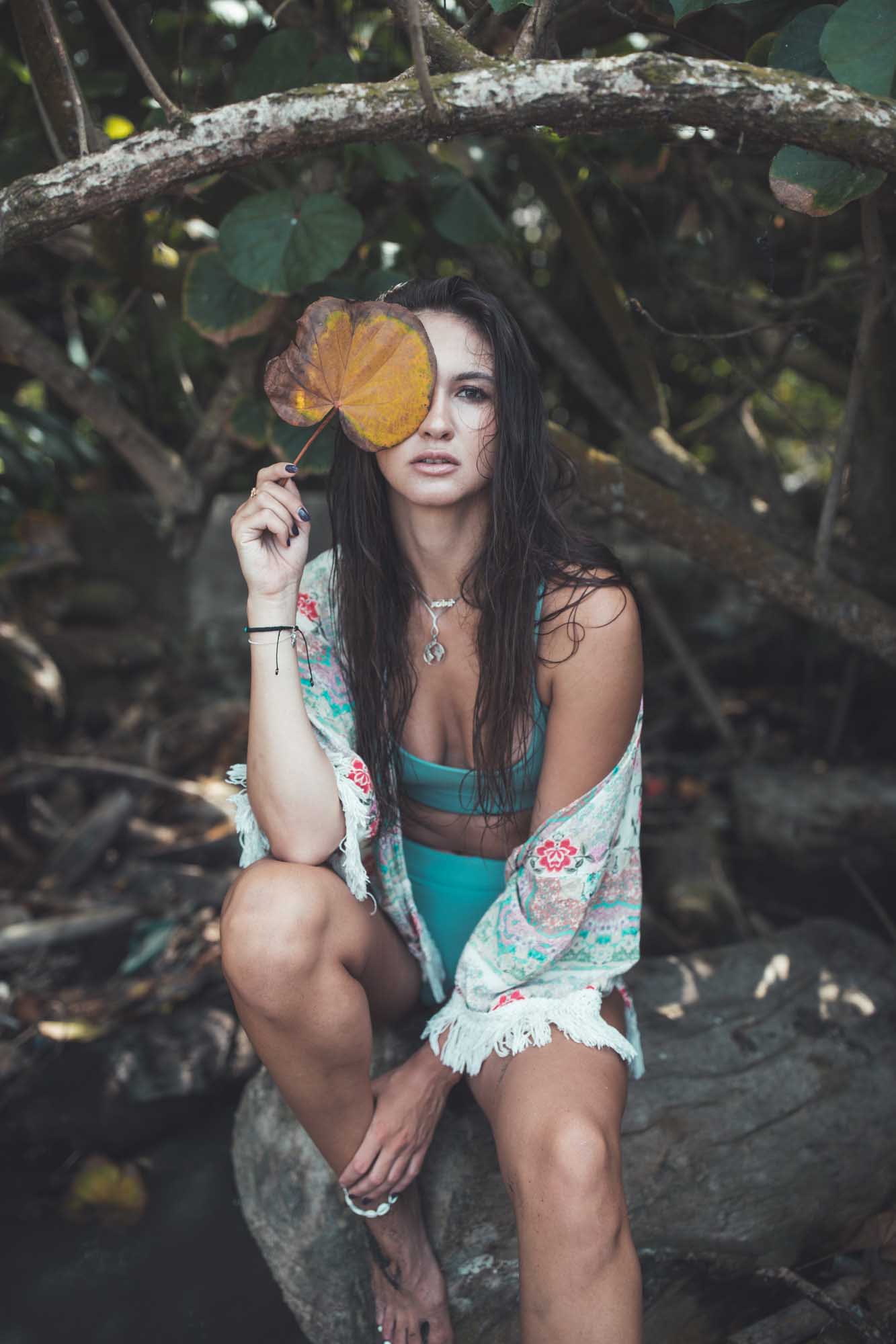 Brunette in a teal bikini and embroidered cover-up crouches on a log in jungle shade, holding a yellow leaf in front of one eye — Pantai Mangening, Bali.