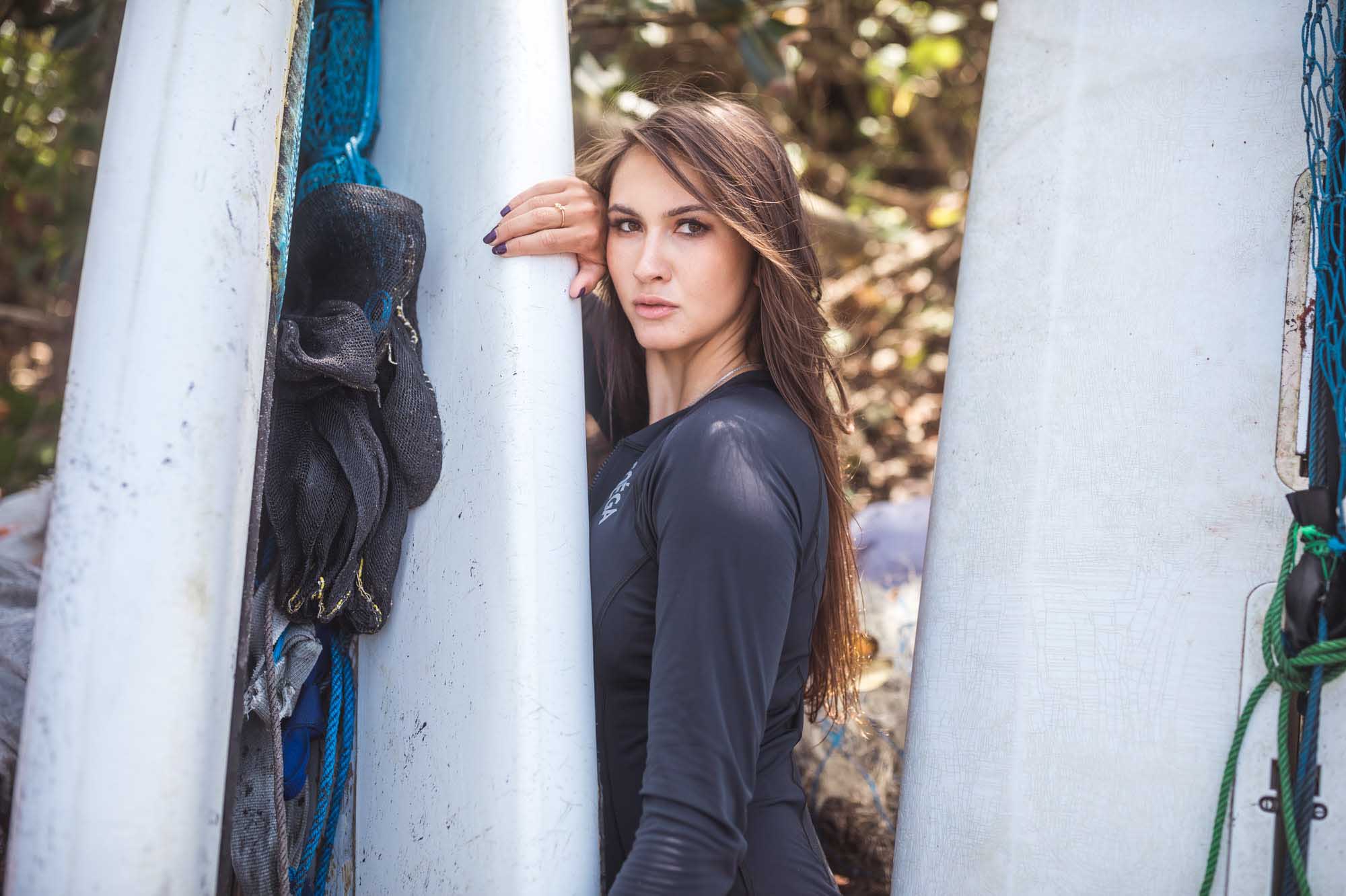 Brunette in a black long-sleeve rashguard leans between stacked white surfboards on a Pantai Mangening beach, Bali.