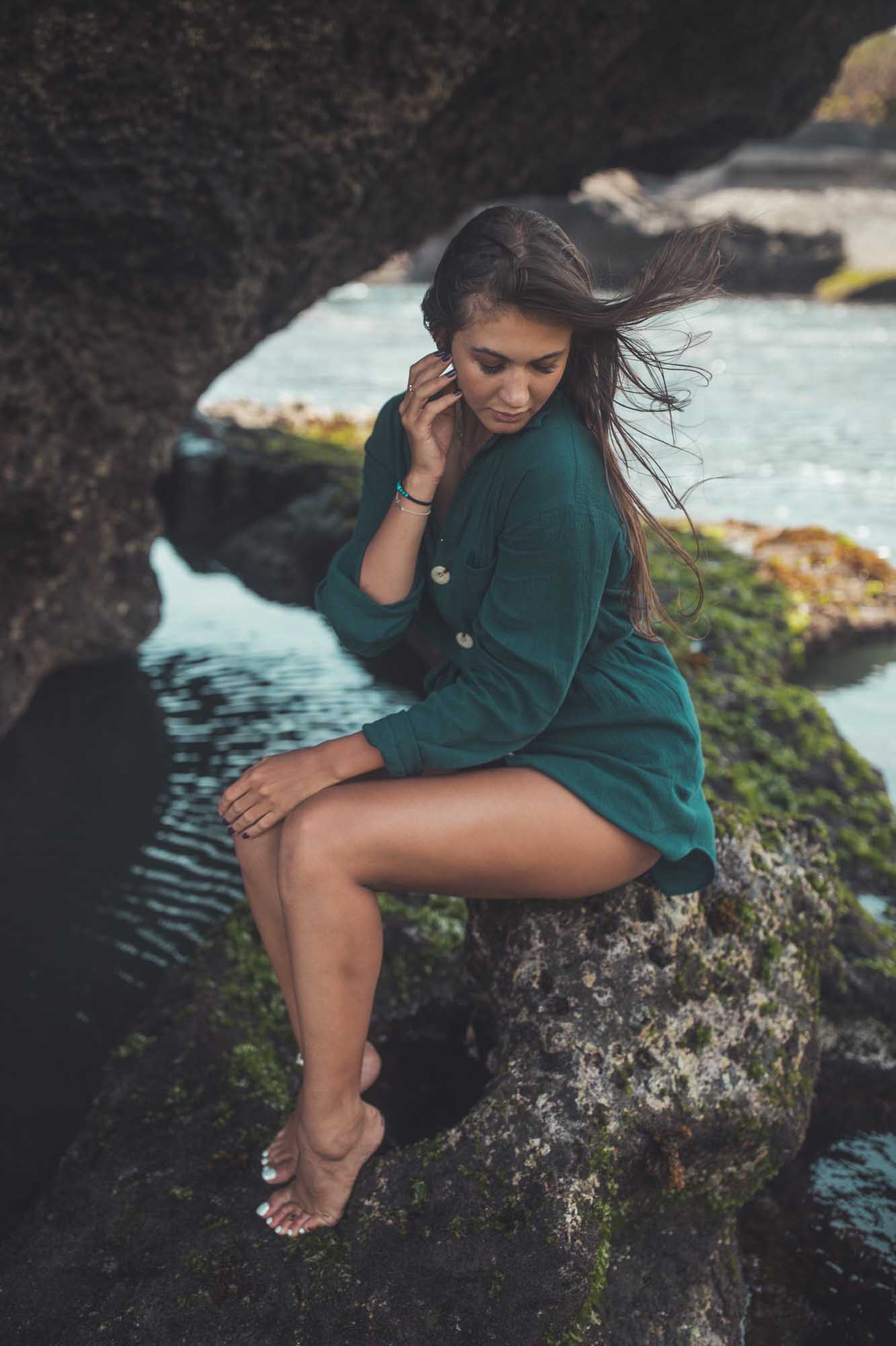 Brunette in a green shirt-dress sits on moss-flecked rocks above a coastal tide pool, hair catching the wind — Pantai Mangening, Bali.