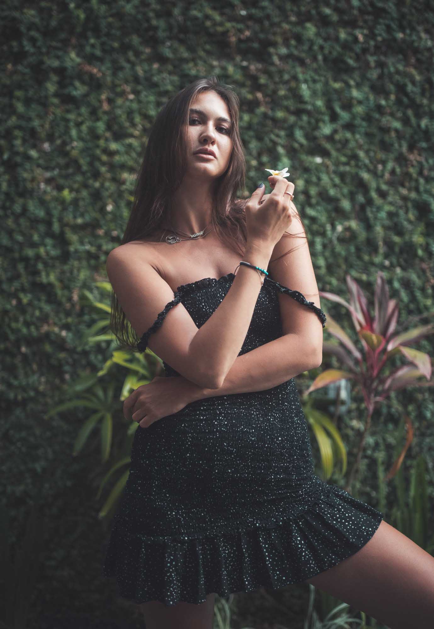 Brunette in a black sequined mini-dress holds a small white frangipani flower against a wall of green ivy — Canggu, Bali.