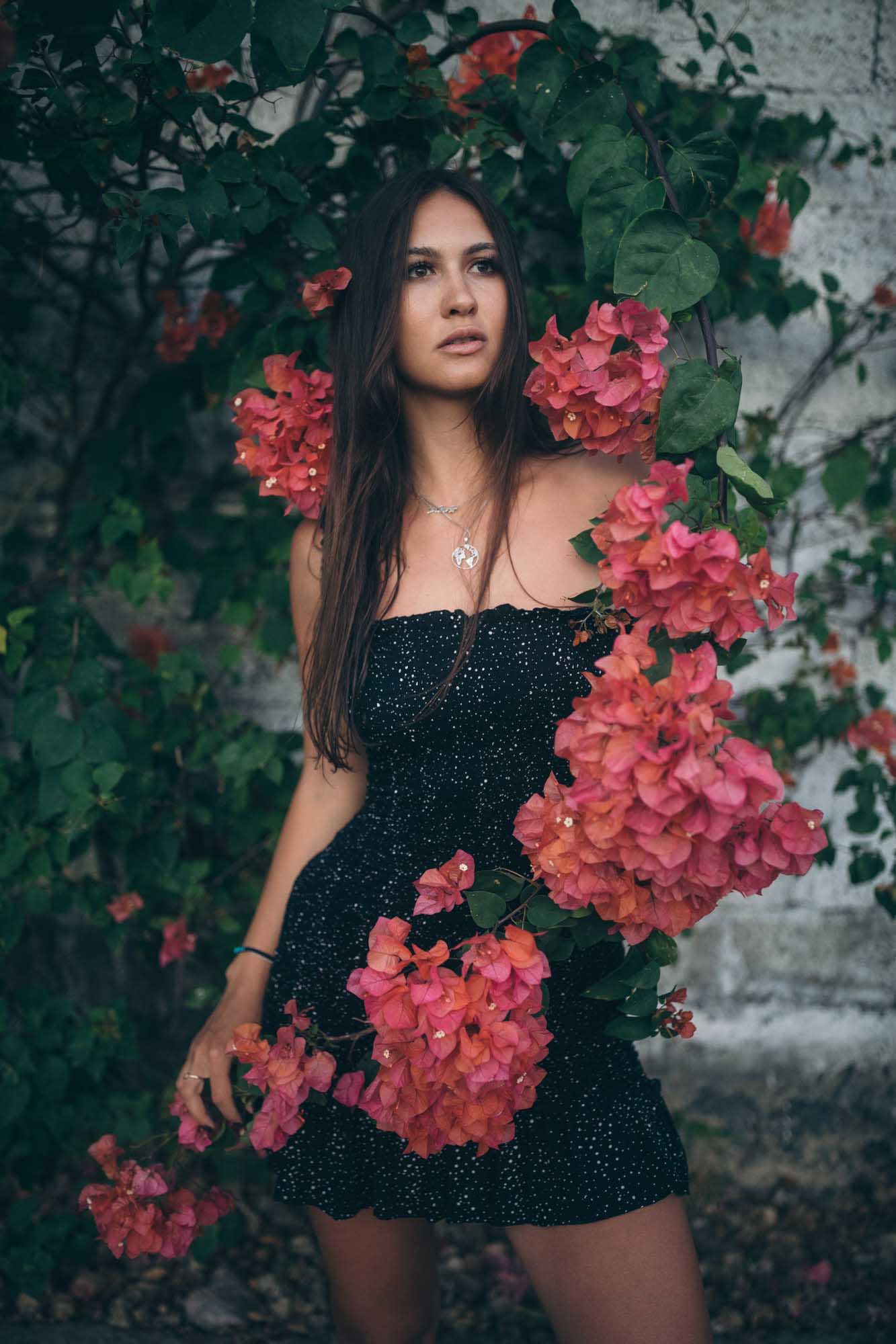 Brunette in a black sequined mini-dress poses beside a cascade of pink bougainvillea on a weathered white wall — Canggu, Bali.