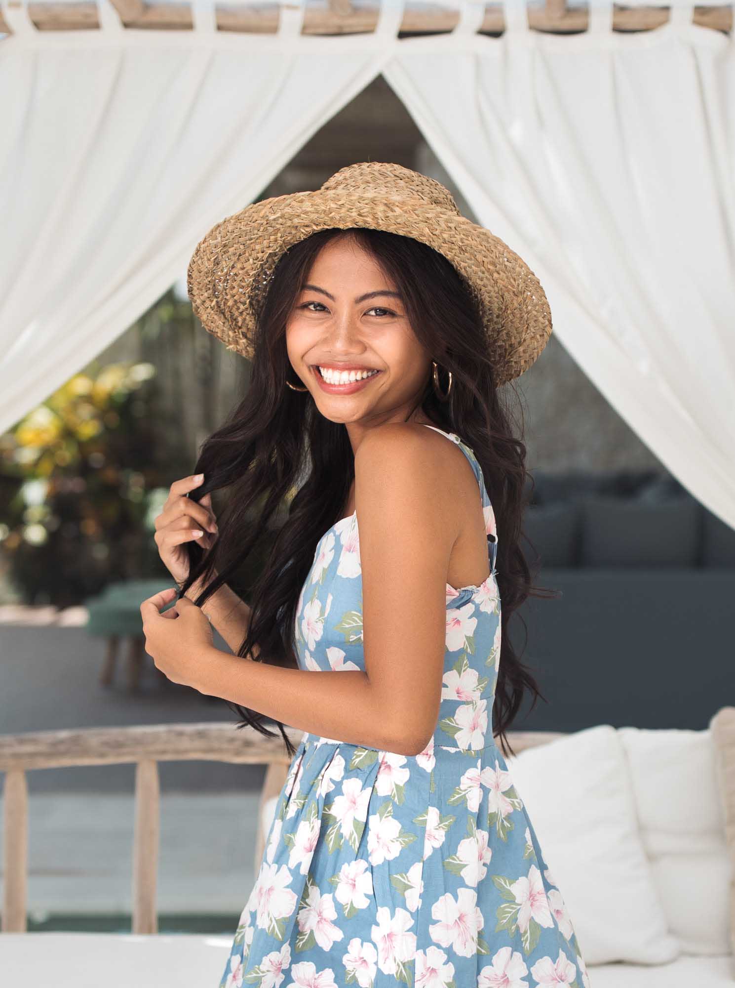 Hailey Ori in a teal hibiscus sundress and straw hat smiles beside the white curtains of a villa daybed — Canggu, Bali.