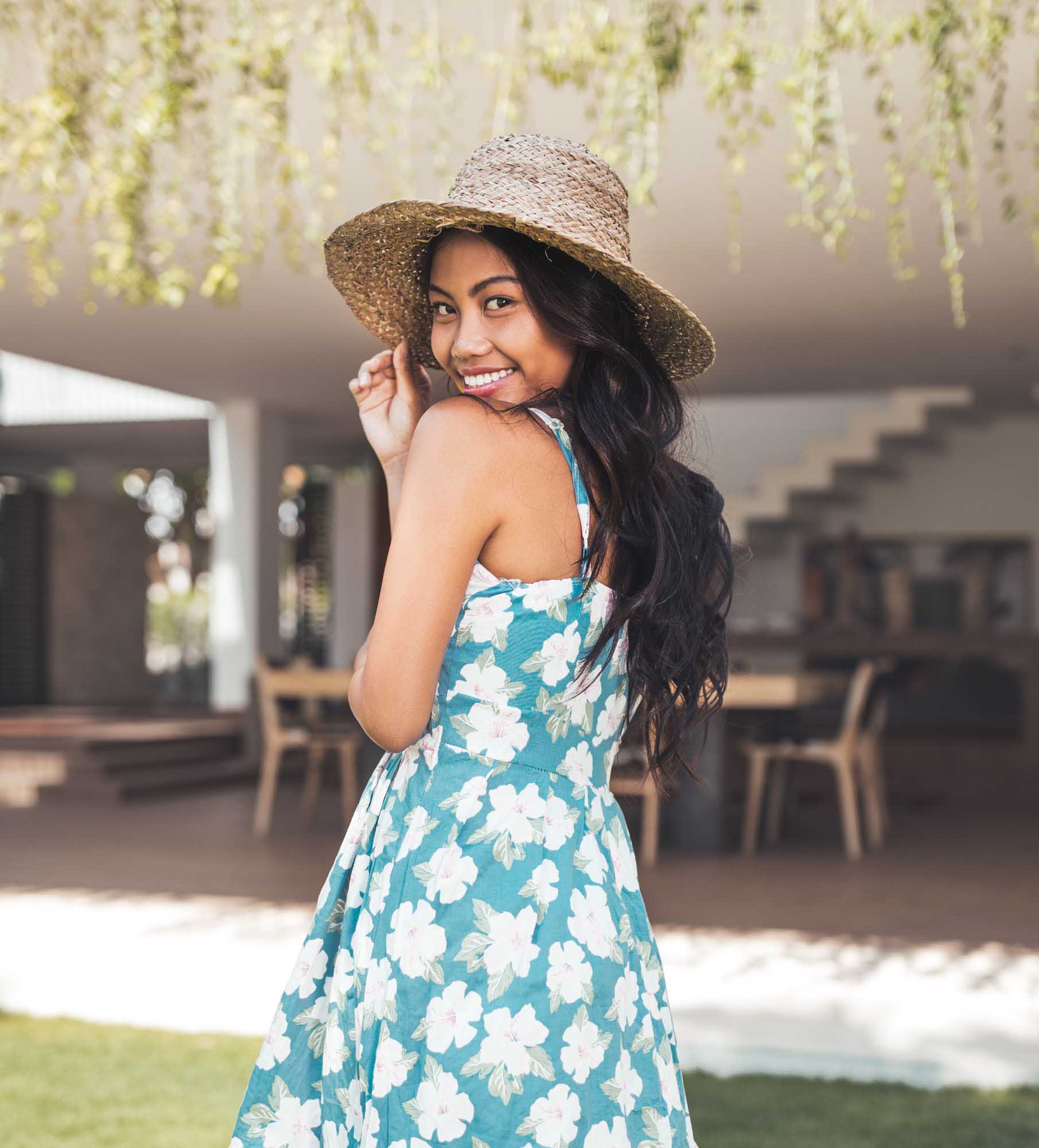 Hailey Ori in a teal hibiscus-print sundress and straw hat smiles over her shoulder on a sunlit villa lawn — Canggu, Bali.