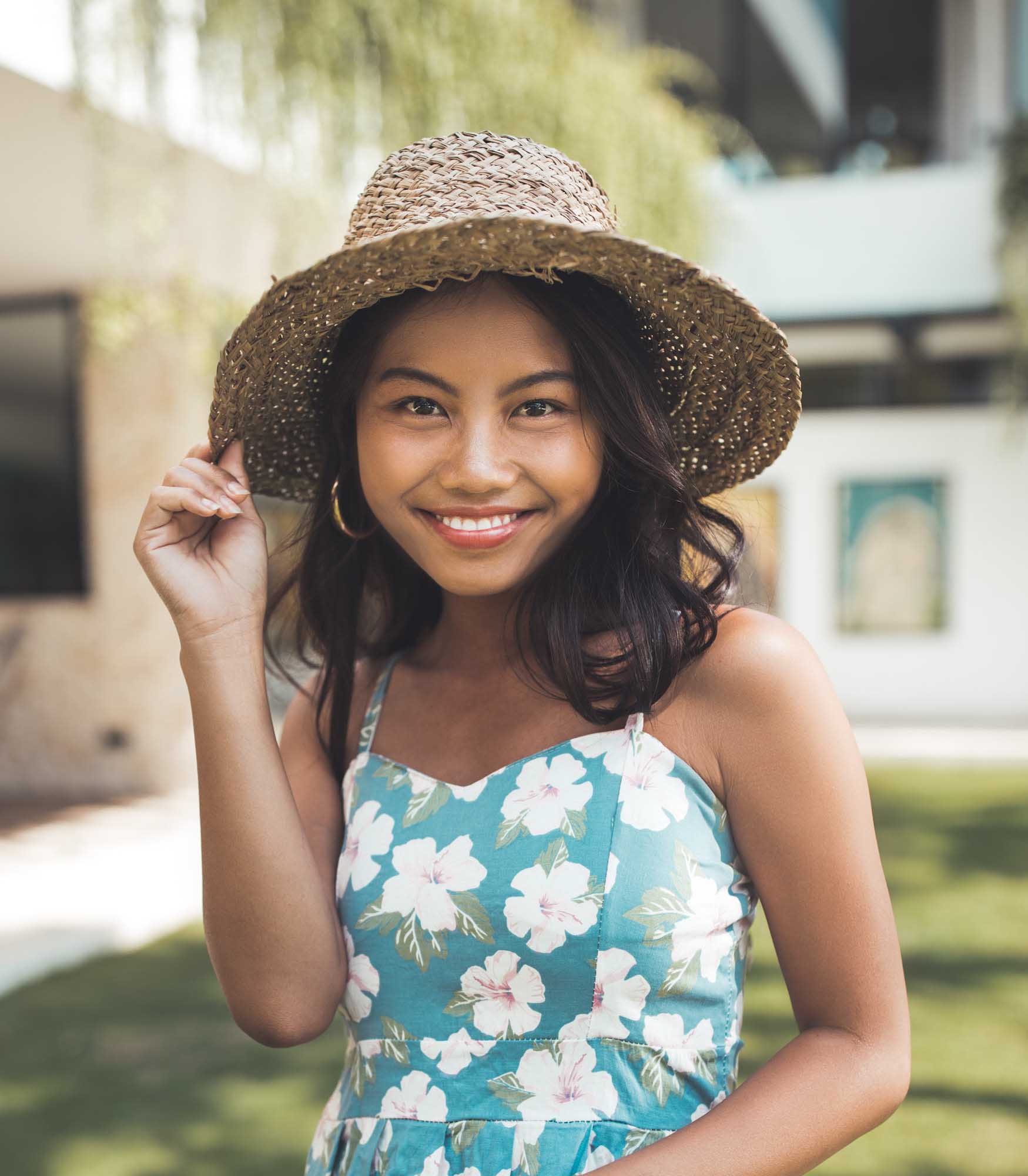 Close-up smile of Hailey Ori in a teal hibiscus sundress and straw hat on a sunlit villa lawn — Canggu, Bali.