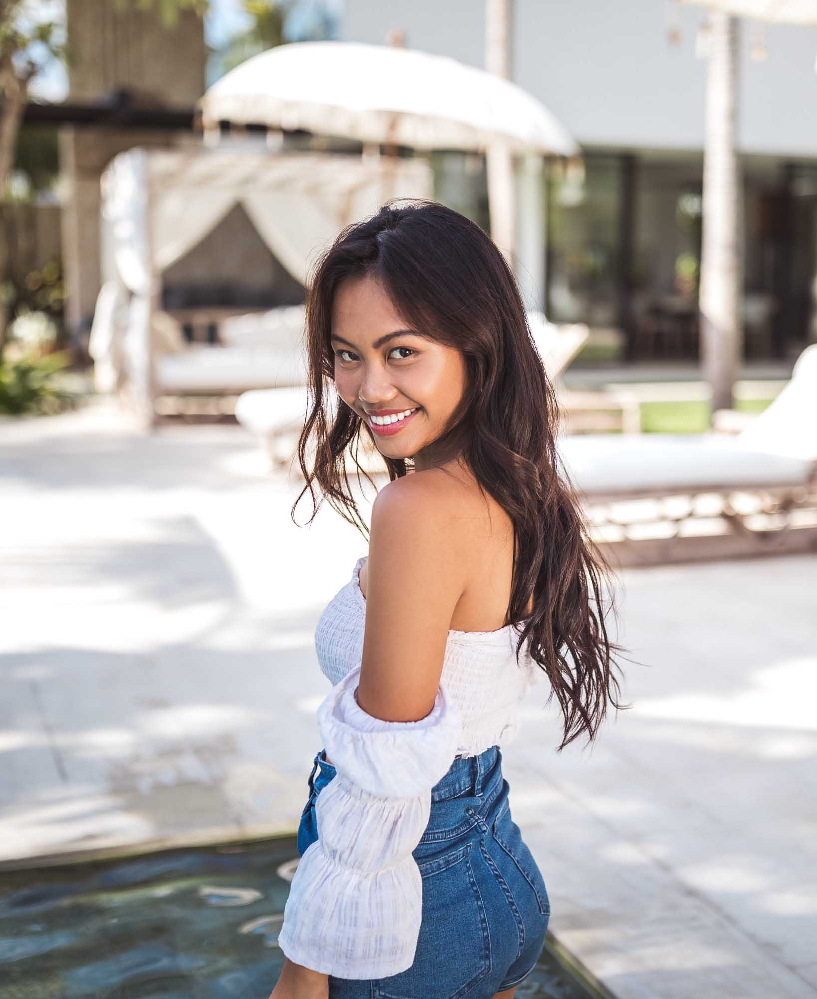 Hailey Ori in a white off-shoulder smocked top and denim shorts smiles over her shoulder on a sunlit villa deck — Canggu, Bali.