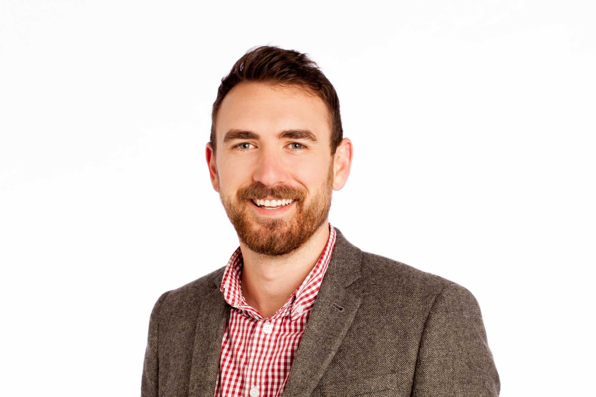 Studio headshot of Alex Guerri on a white background — brunette with a red beard, grey tweed blazer over a red-and-white gingham button-down.