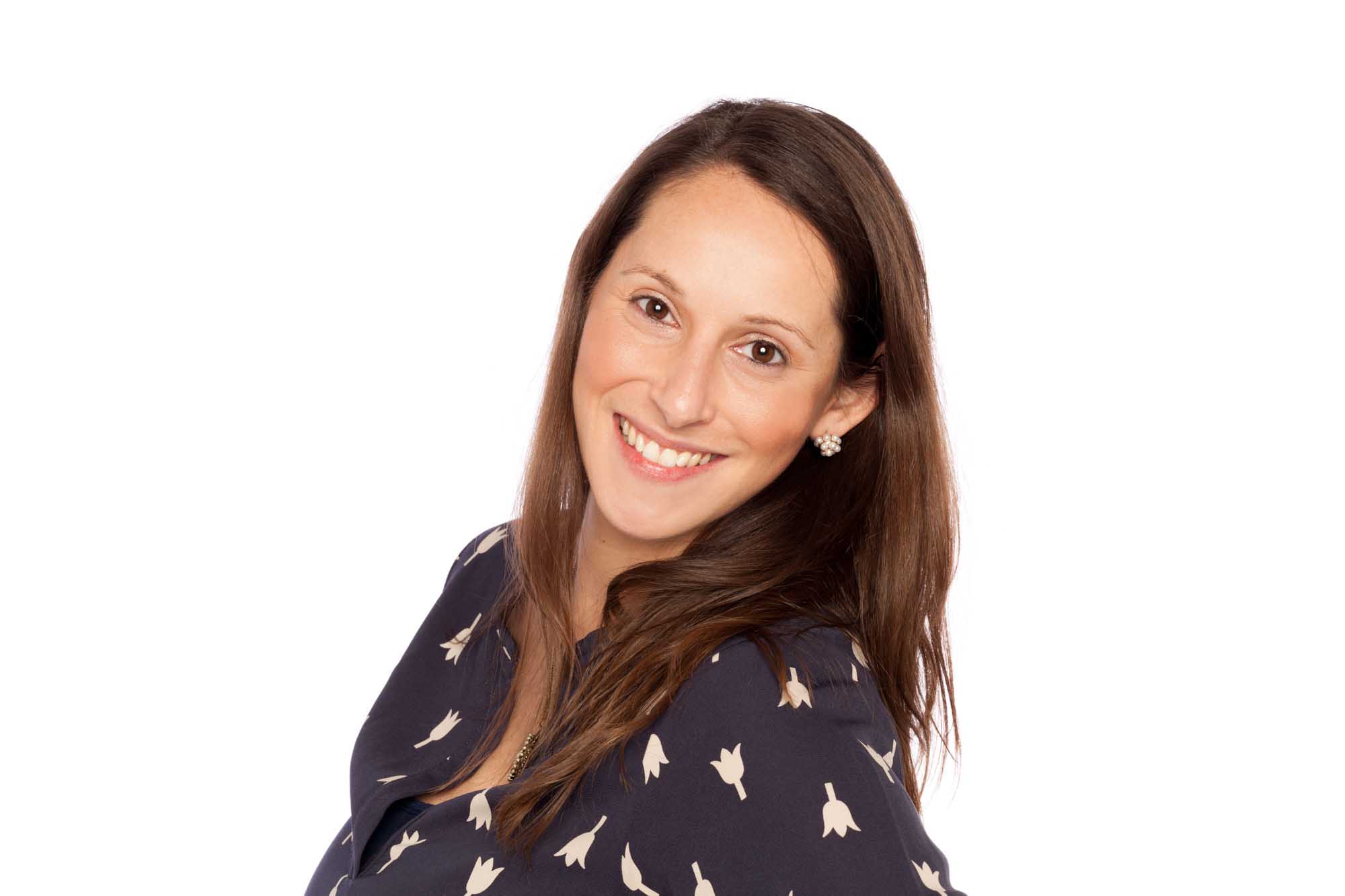 Studio headshot of Anne Catherine Feeney on a white background — long brown hair, warm smile, navy blouse with a printed motif.