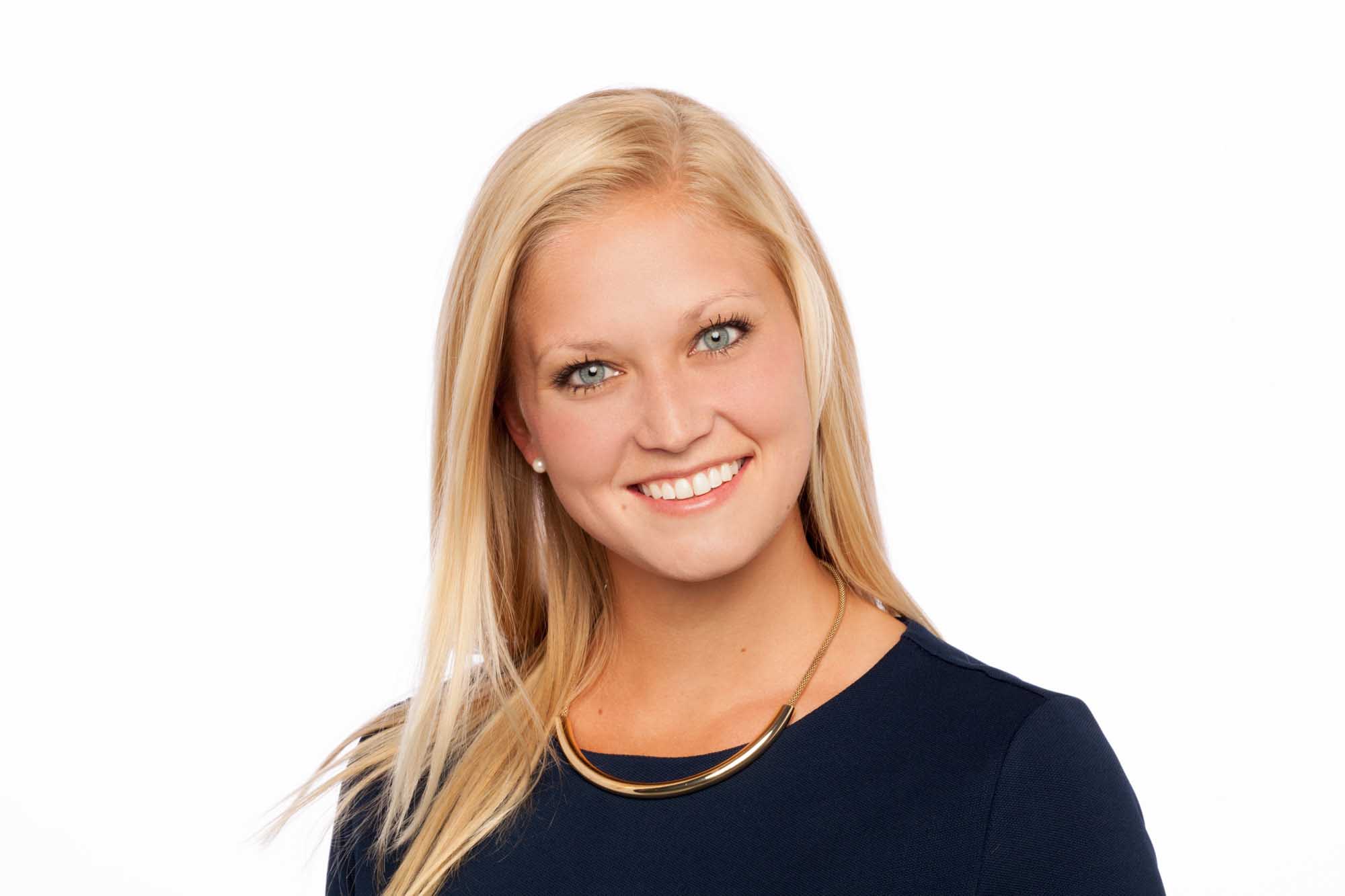 Studio headshot of Ashley Huehnerfuss on a white background — long blonde hair, warm smile, dark navy top and chunky gold collar necklace.