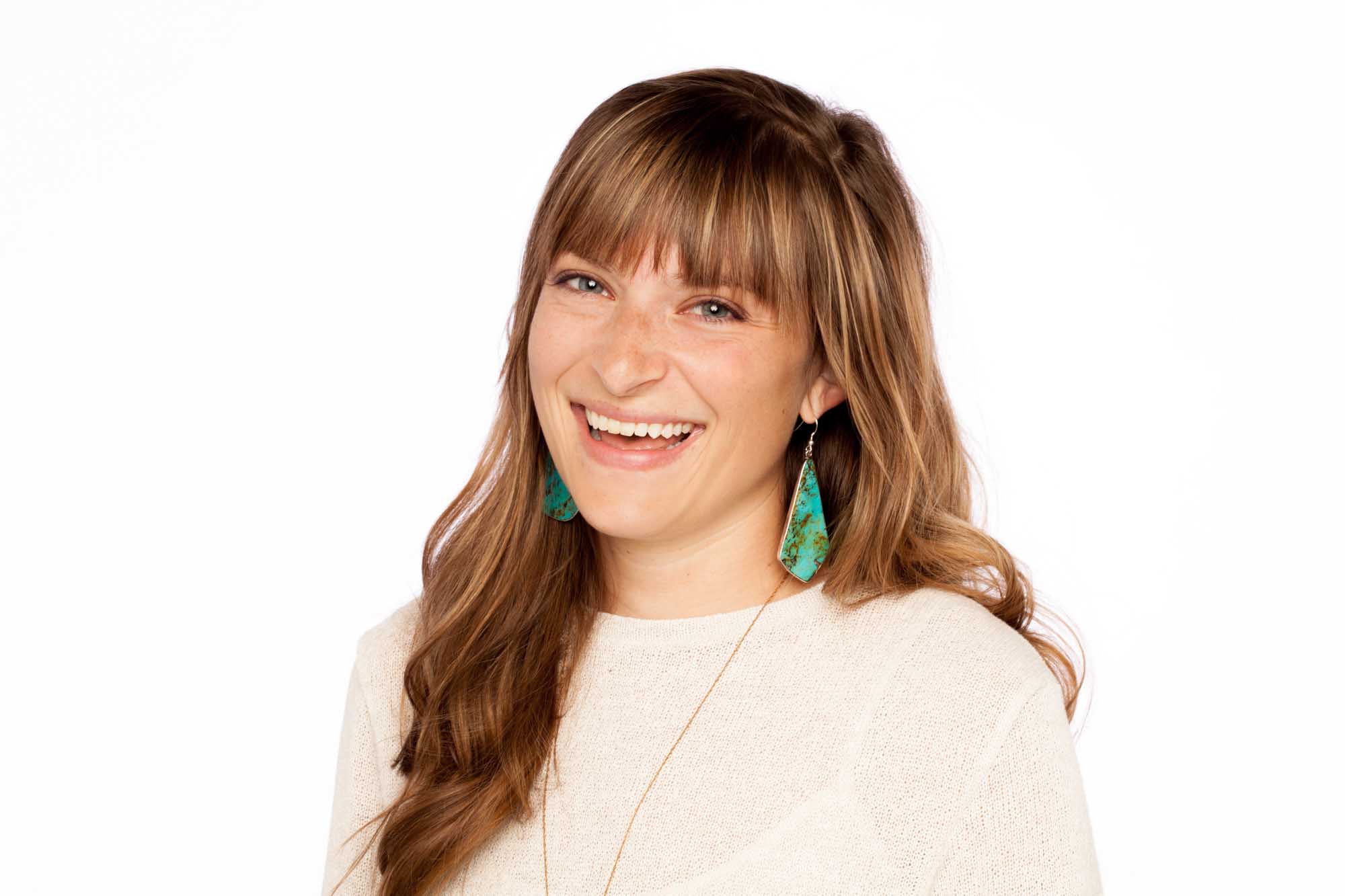 Studio headshot of Bethany Lechner on a white background — wavy brown hair with bangs, cream textured sweater, turquoise statement earring.