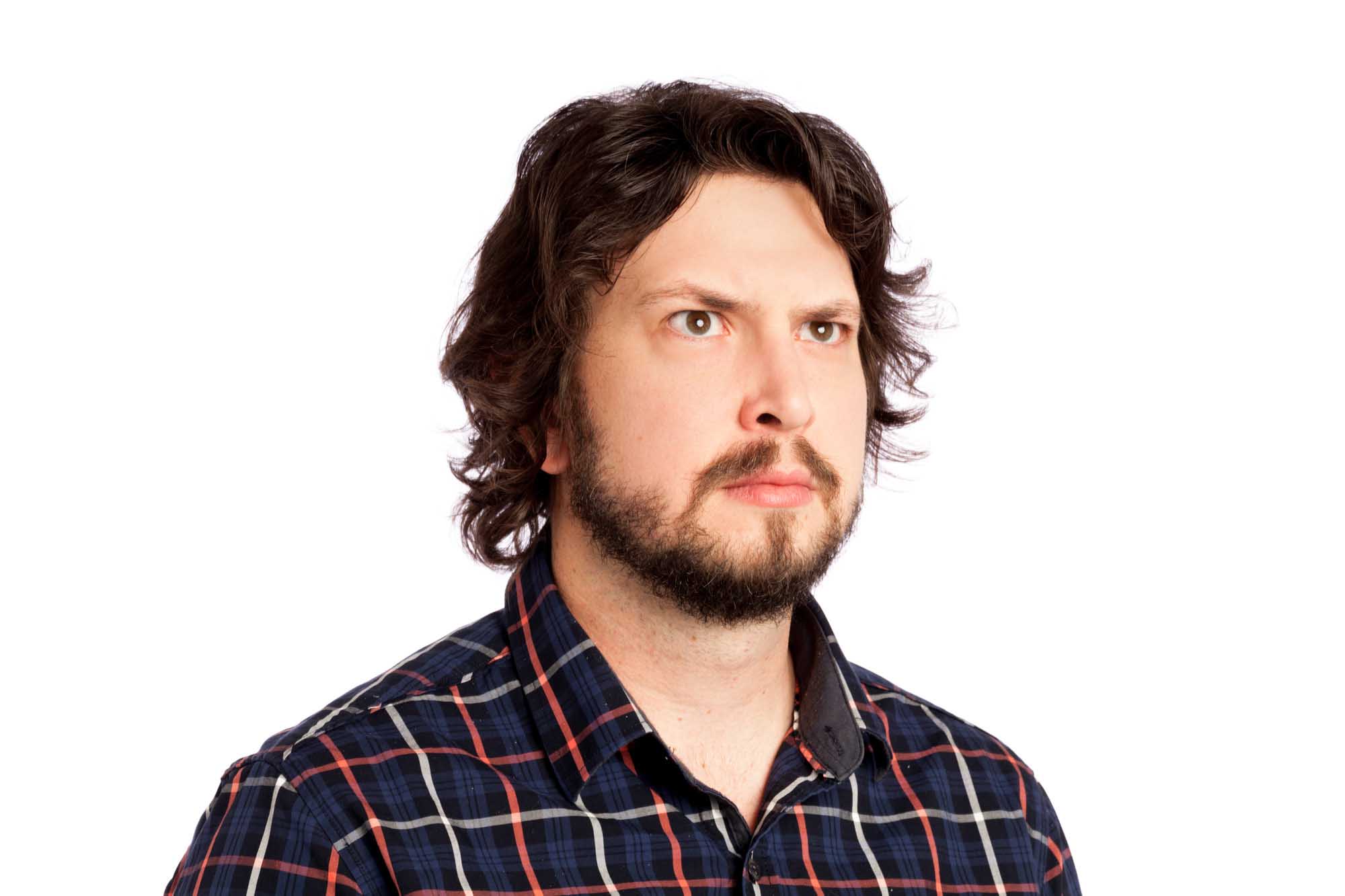 Studio headshot of Dave Rowley on a white background — dark wavy hair, full beard, plaid flannel button-down, deadpan expression.