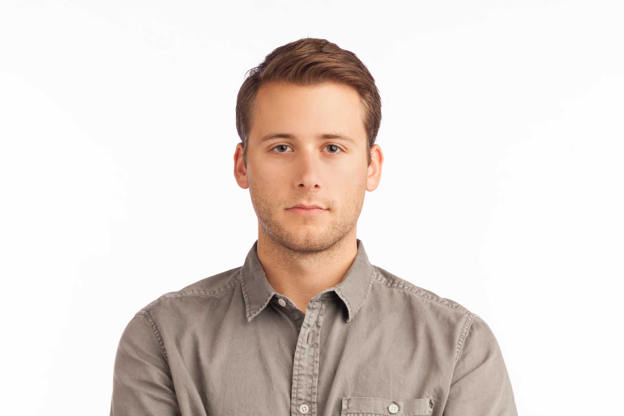 Studio headshot of Dominic Scariano on a white background — short brown hair, light stubble, grey button-down shirt.