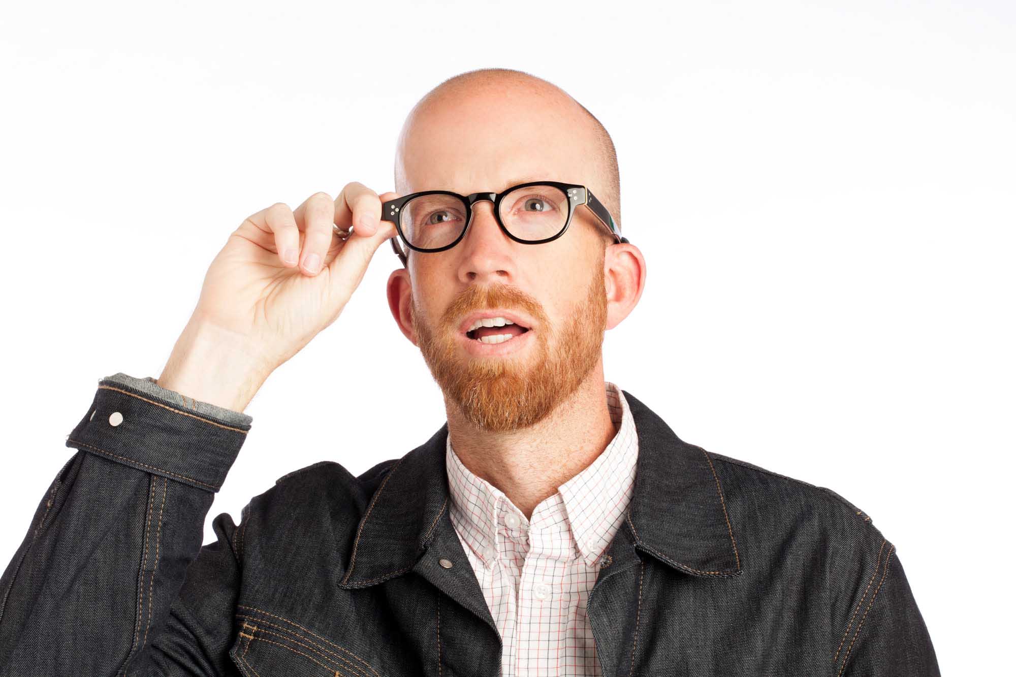 Studio headshot of Josh Shelton on a white background — bald, red beard, black-framed glasses, denim jacket over a pink gingham shirt.