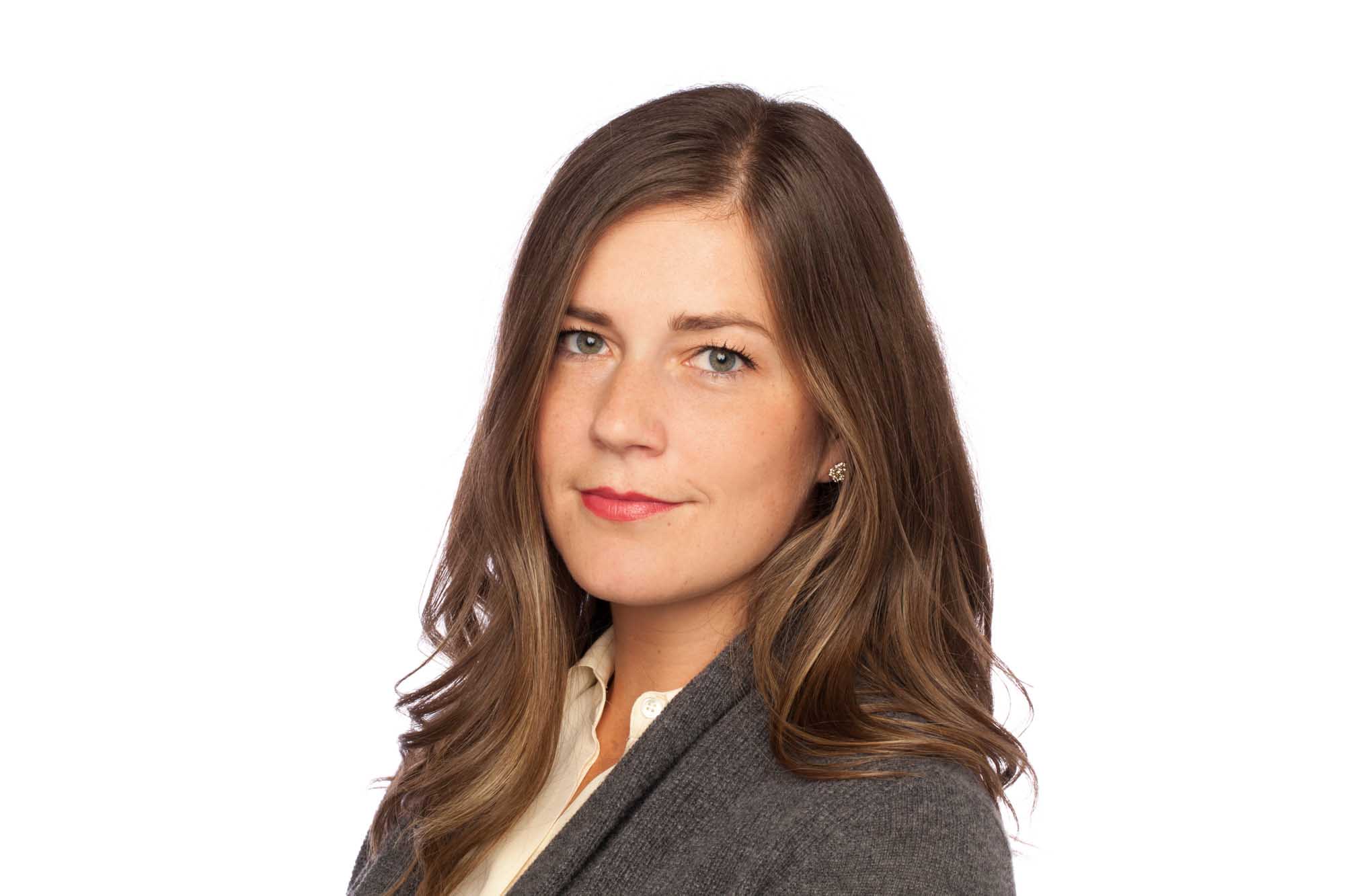 Studio headshot of Lindsey Melloh on a white background — long brunette hair, grey blazer over a cream blouse, composed expression.