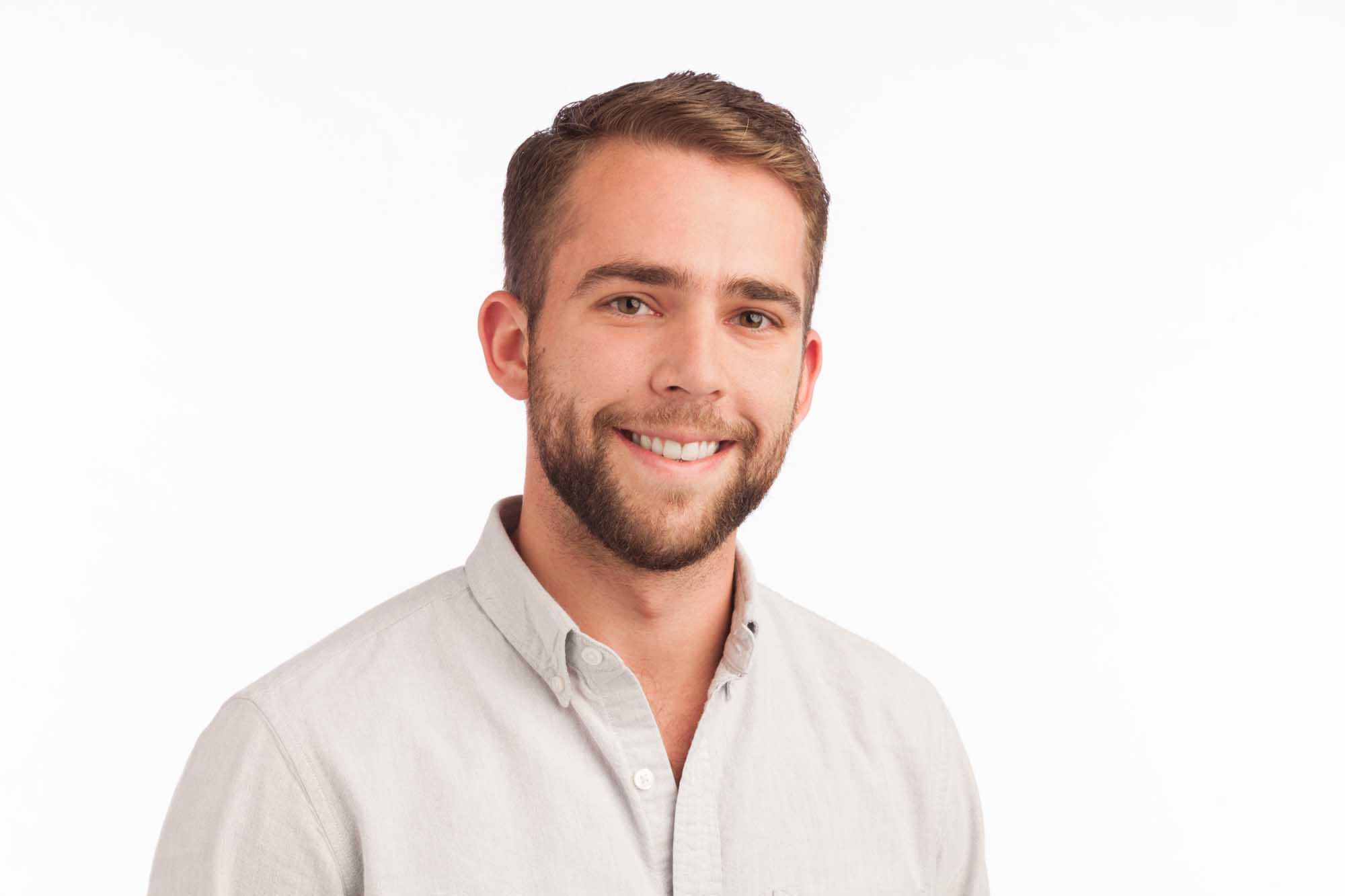 Studio headshot of Sam Carolan on a white background — short brown hair, full beard, light-grey oxford button-down, warm smile.
