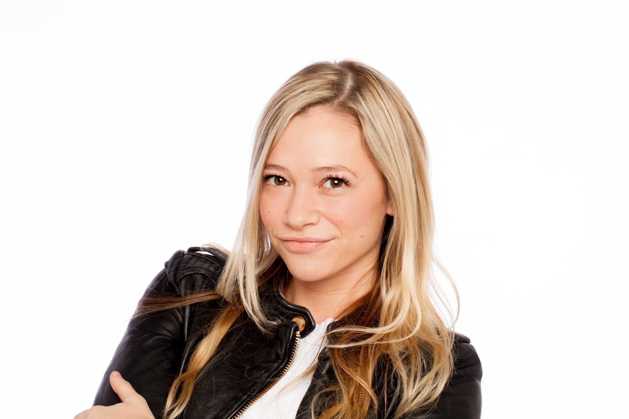 Studio headshot of Sarah Stuchbery on a white background — layered blonde hair, black leather jacket over a white tee, arms crossed, smiling.