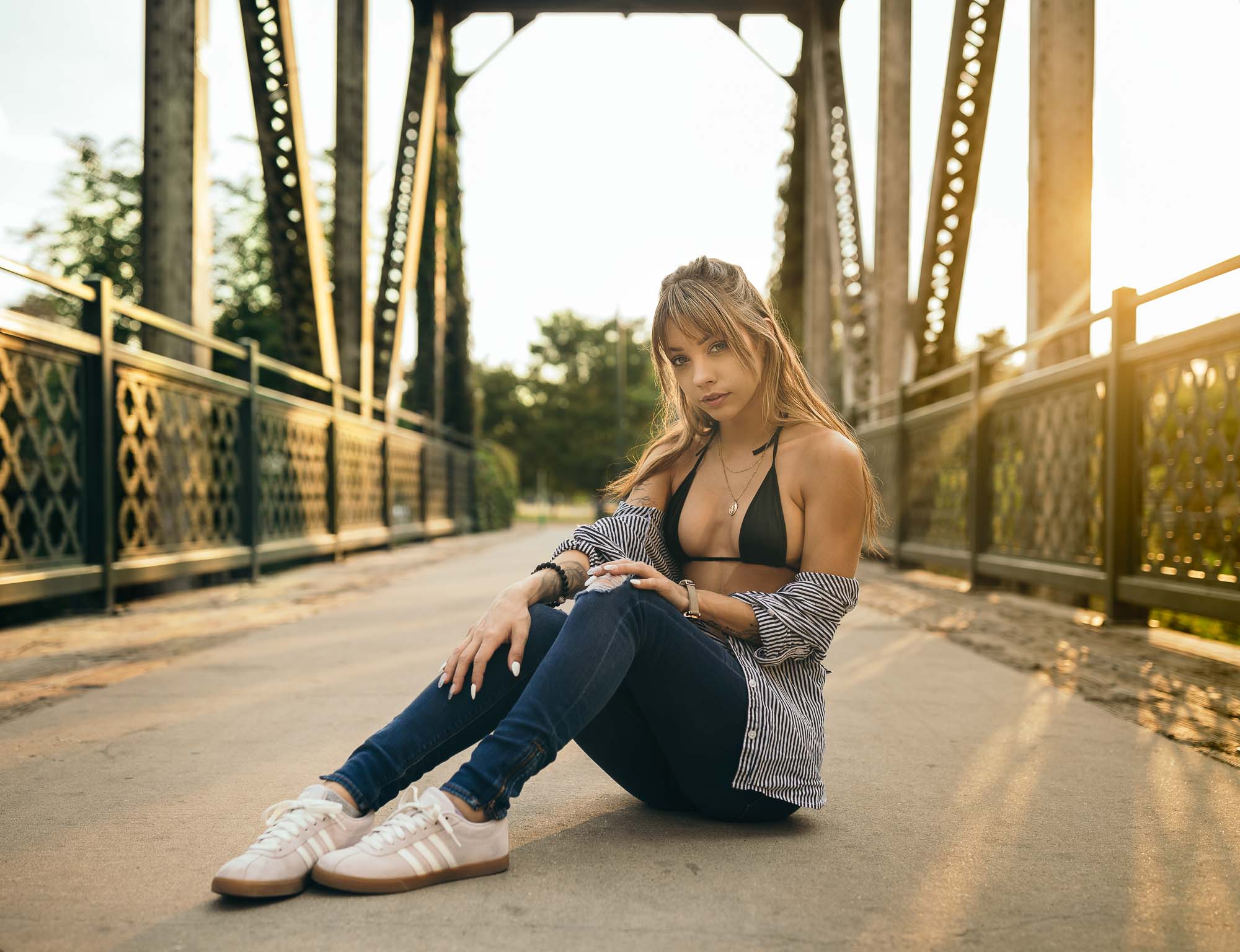 Model @space.junkiee sits cross-legged on the walkway of a steel-truss pedestrian bridge at golden hour, wearing a black bikini top under an open striped shirt, jeans and white sneakers.
