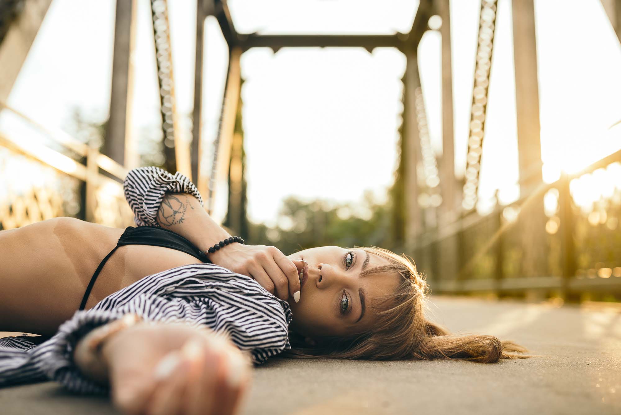 Editorial fashion photo of model @space.junkiee lying on a Denver bridge at sunset wearing a striped shirt