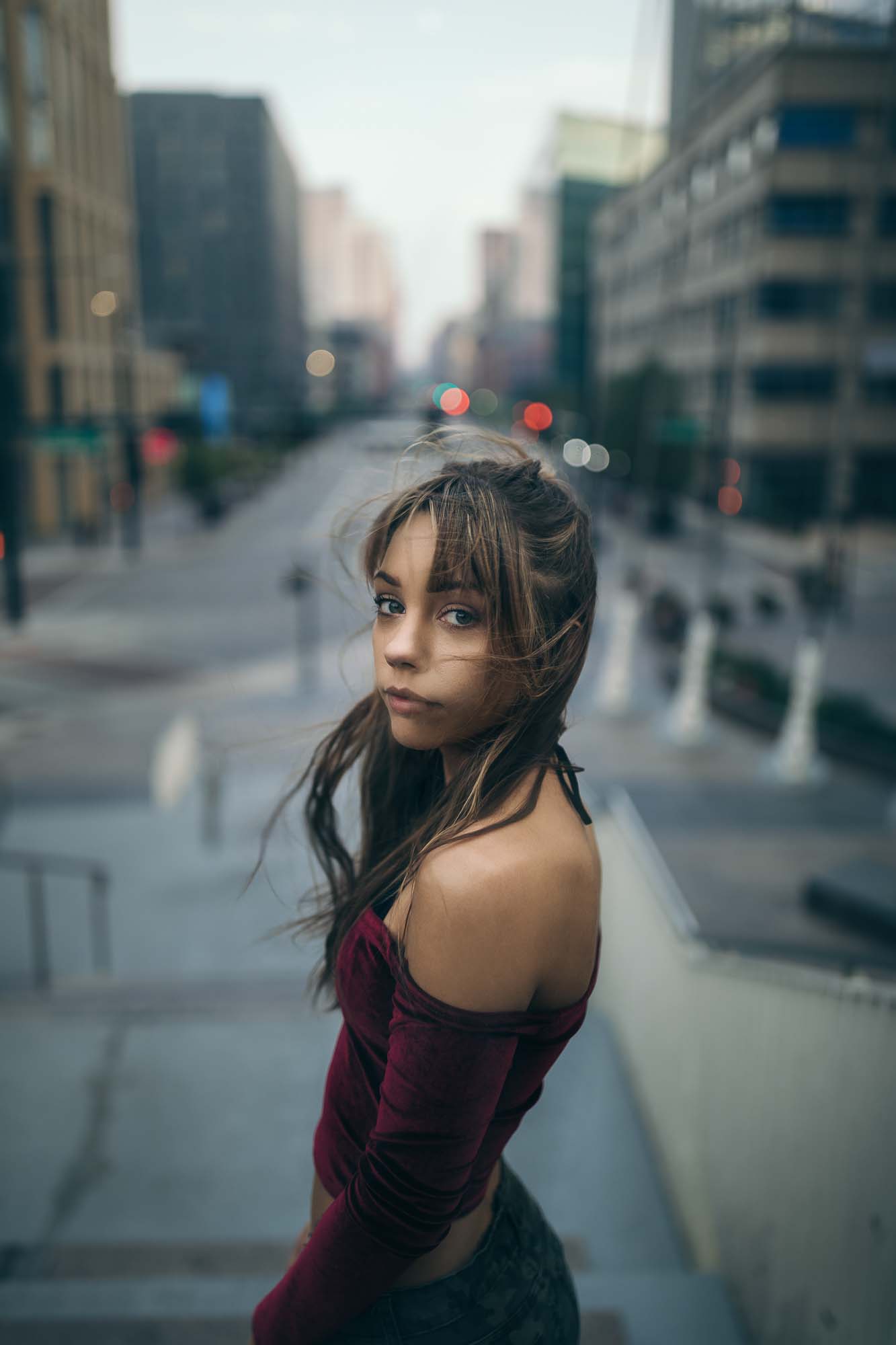 Editorial portrait of model @space.junkiee on stairs in downtown Denver at dusk with city light bokeh