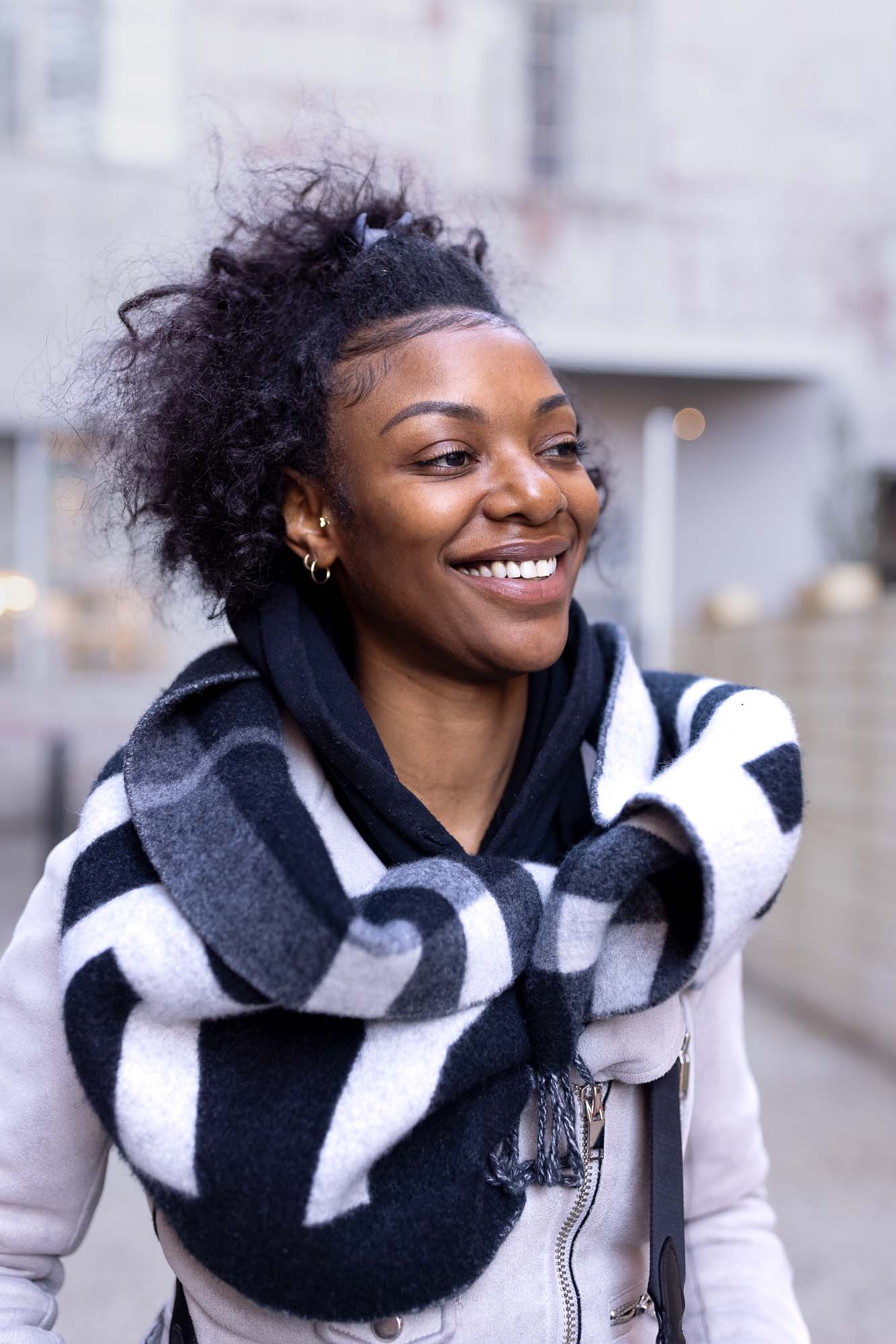 Candid street-style portrait of a woman smiling and looking off-camera in a downtown Denver alleyway, wearing a cream leather jacket and an oversized black-and-white wool scarf.