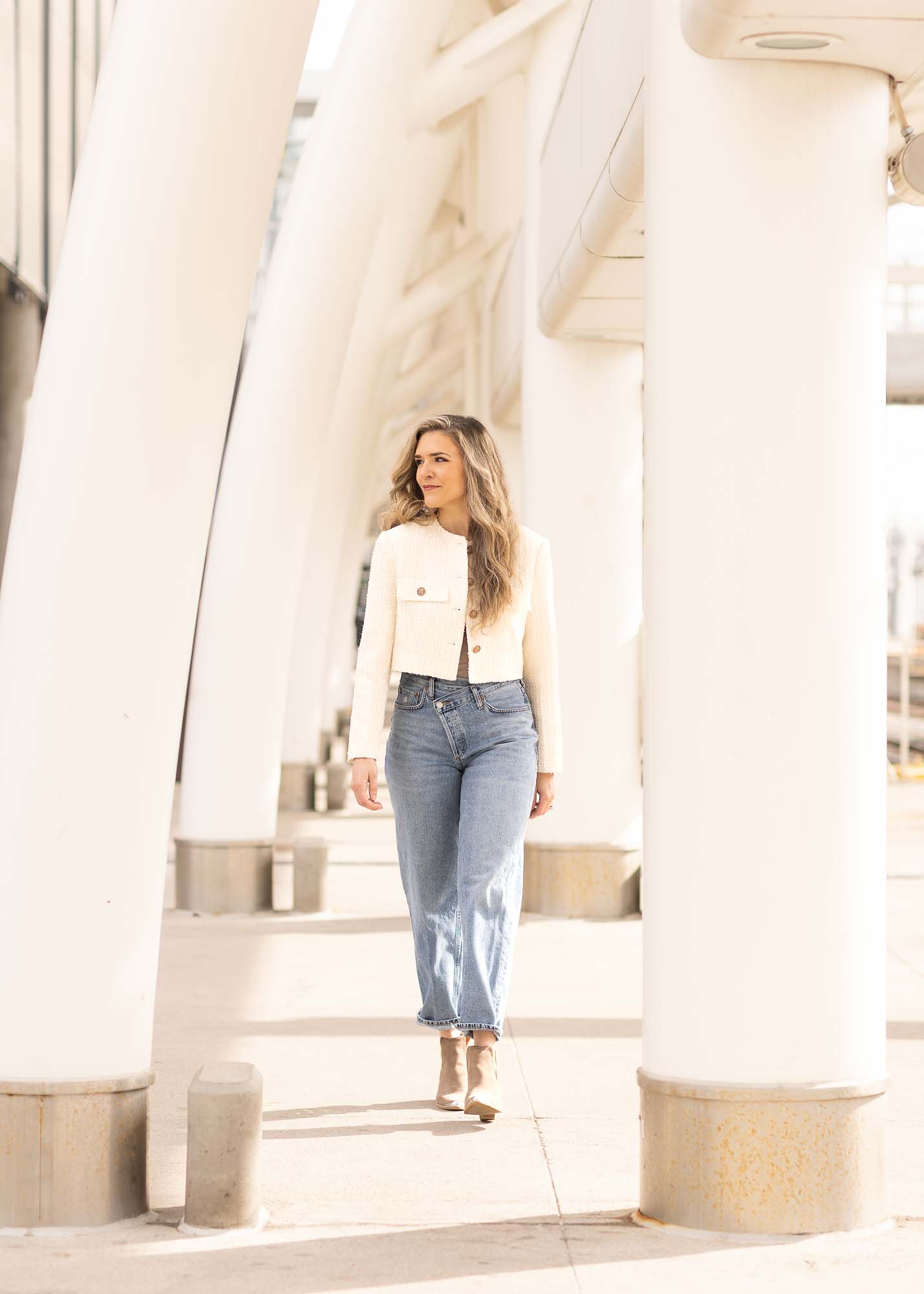 Lifestyle portrait of a woman in a cream cropped jacket and blue jeans walking between the white sculptural columns of the Denver Millennium Bridge.