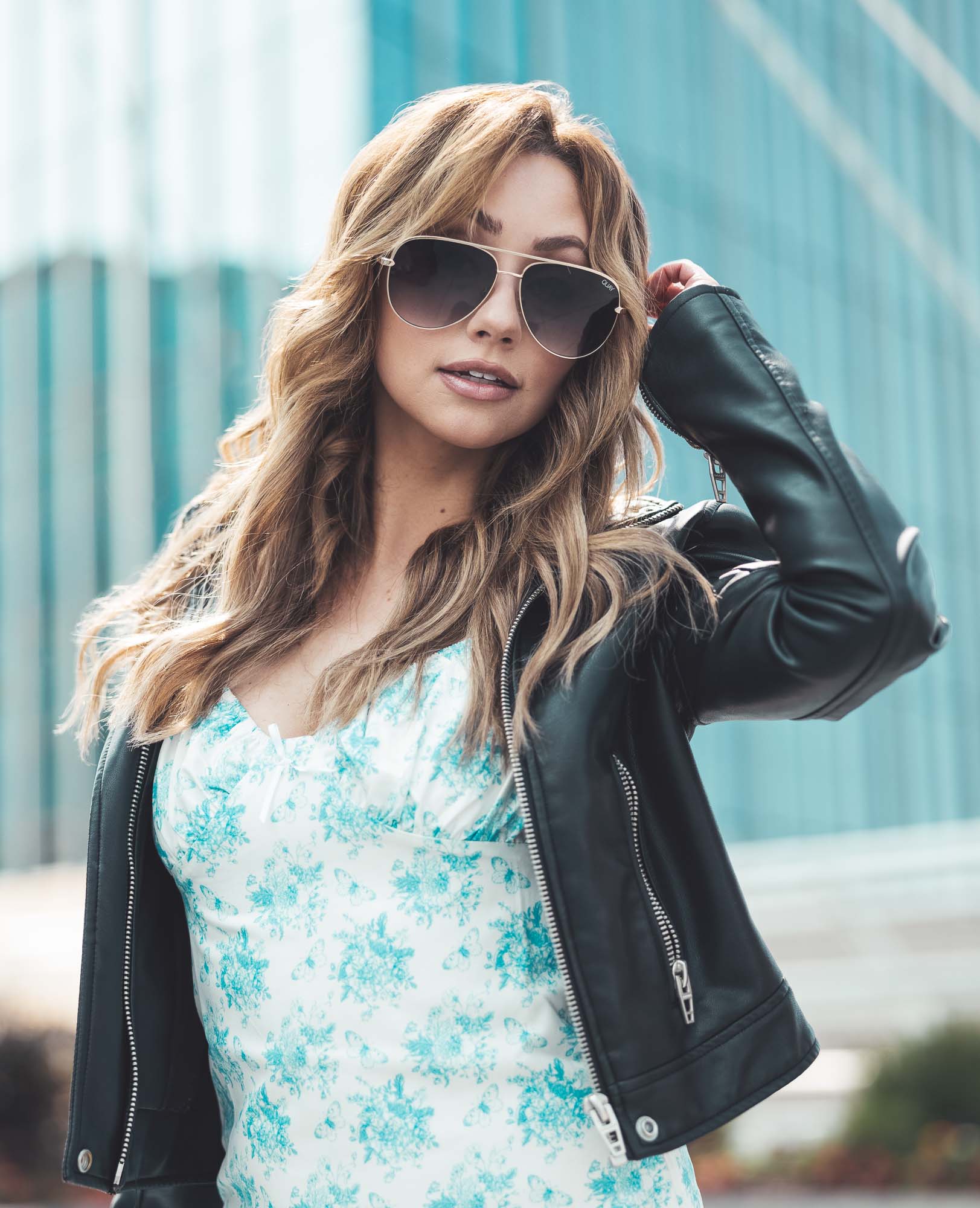 Sara Shimrs in a teal-floral dress, black leather jacket and aviator sunglasses poses against a modern Denver glass facade.