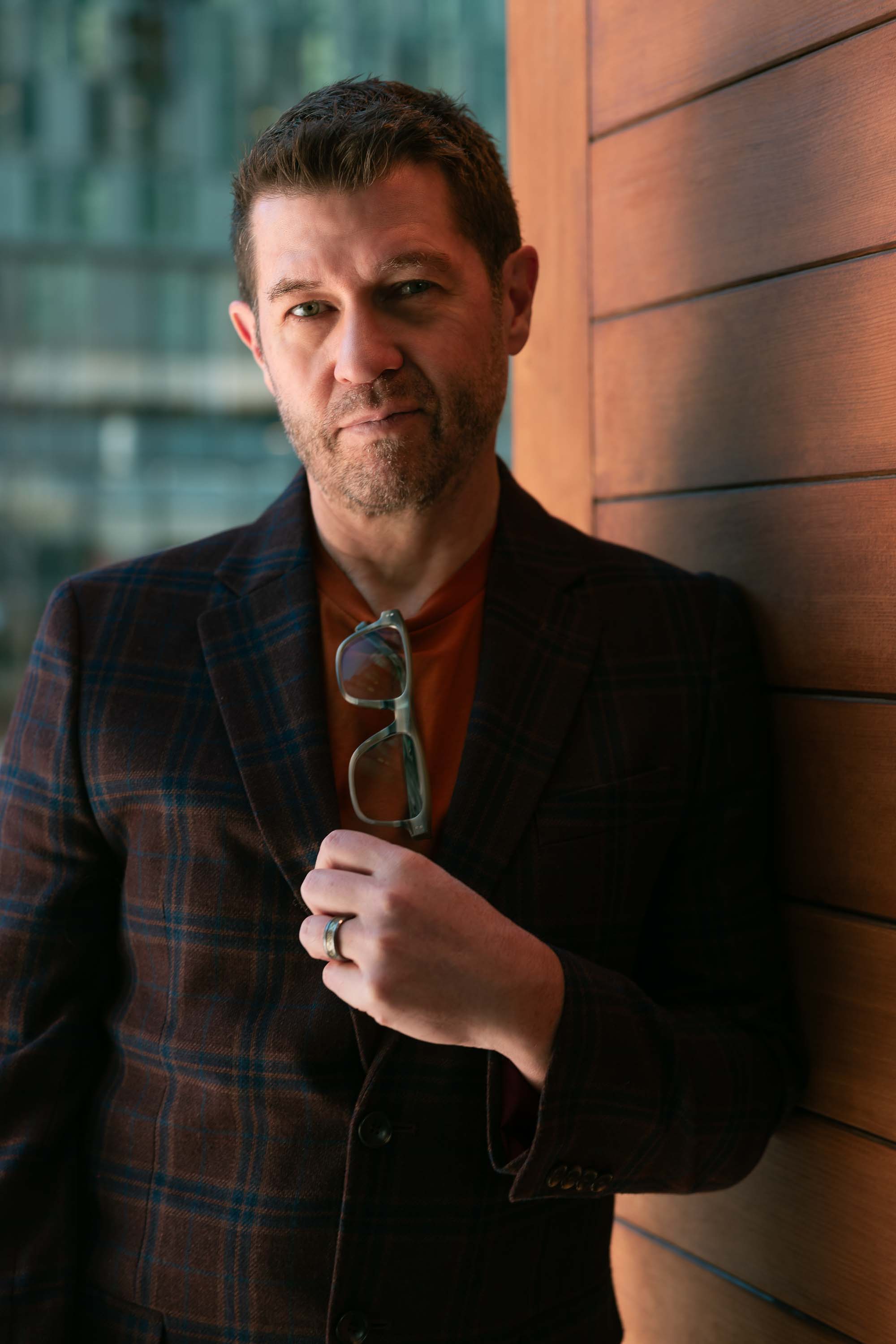 Personal branding portrait of a man in a rust shirt and plaid blazer leaning against a cedar-slat wall in downtown Denver, holding his tortoiseshell glasses.