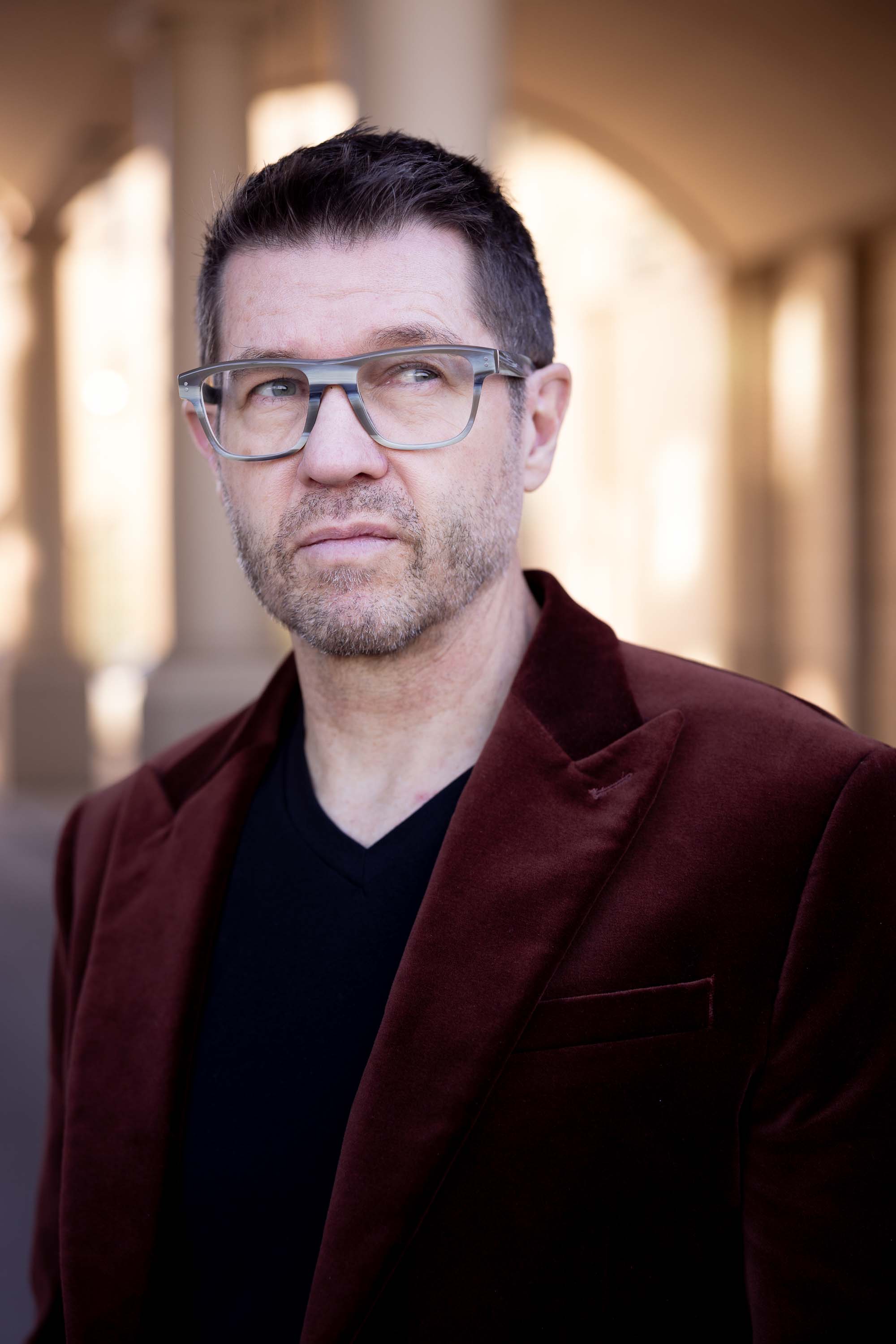 Personal branding portrait of a man in a dark red velvet blazer and glasses standing under an arched Denver colonnade.