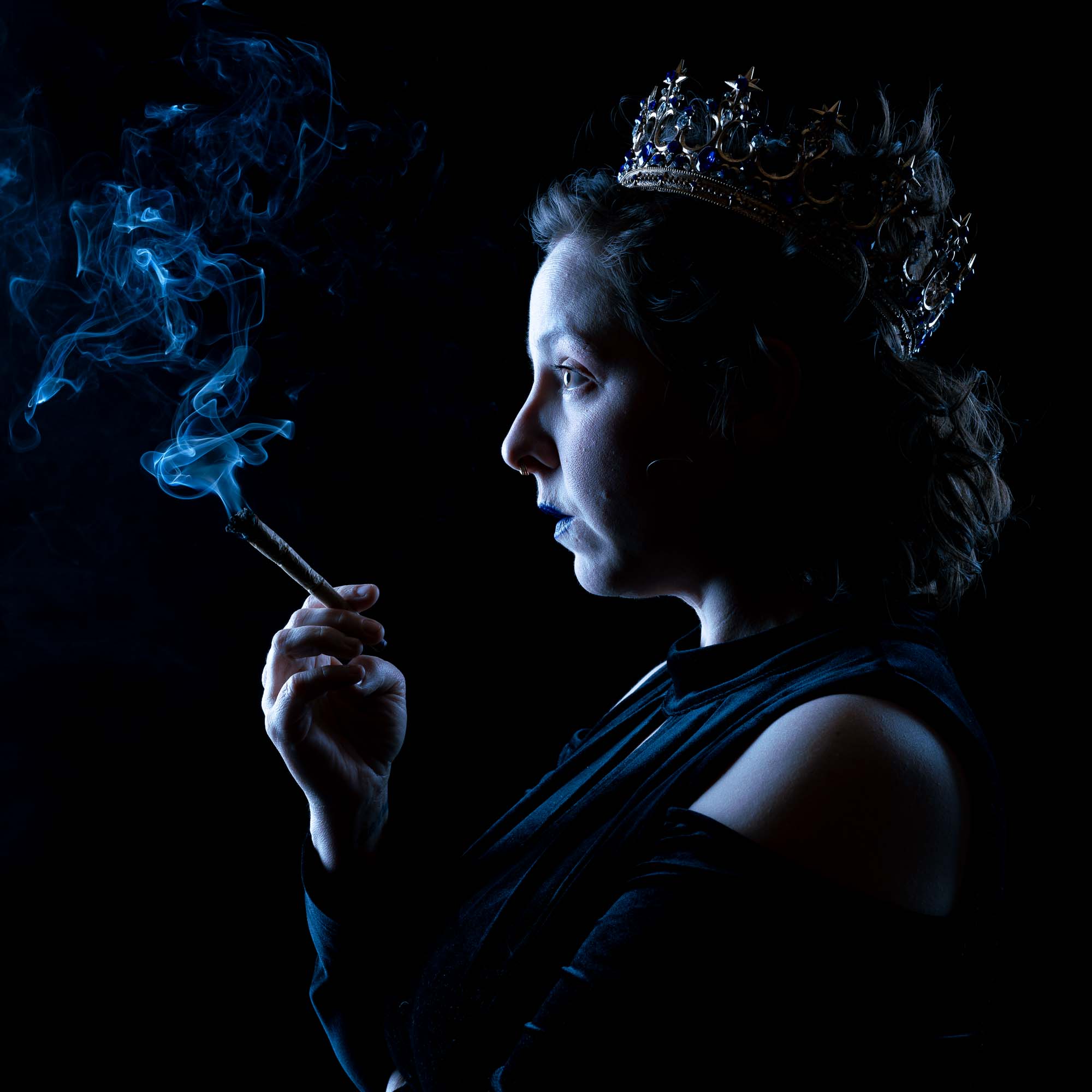 Low-key theatrical portrait of a woman in a crown exhaling smoke in profile, promotional still for Bowls with the Bard.