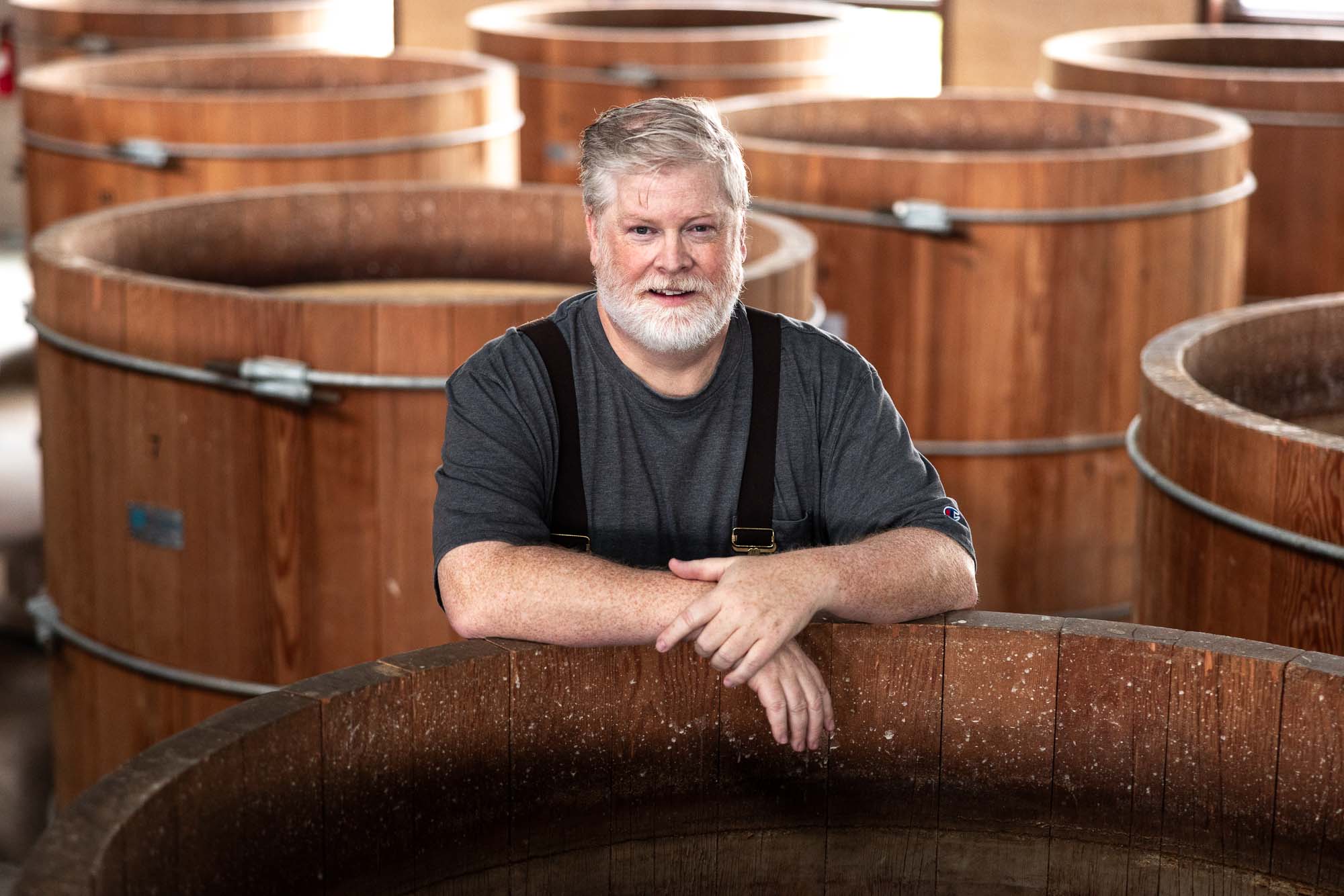Portrait of Todd Leopold, co-founder of Leopold Brothers Distillery, leaning on the rim of a large open-top wooden fermentation vat and smiling at camera