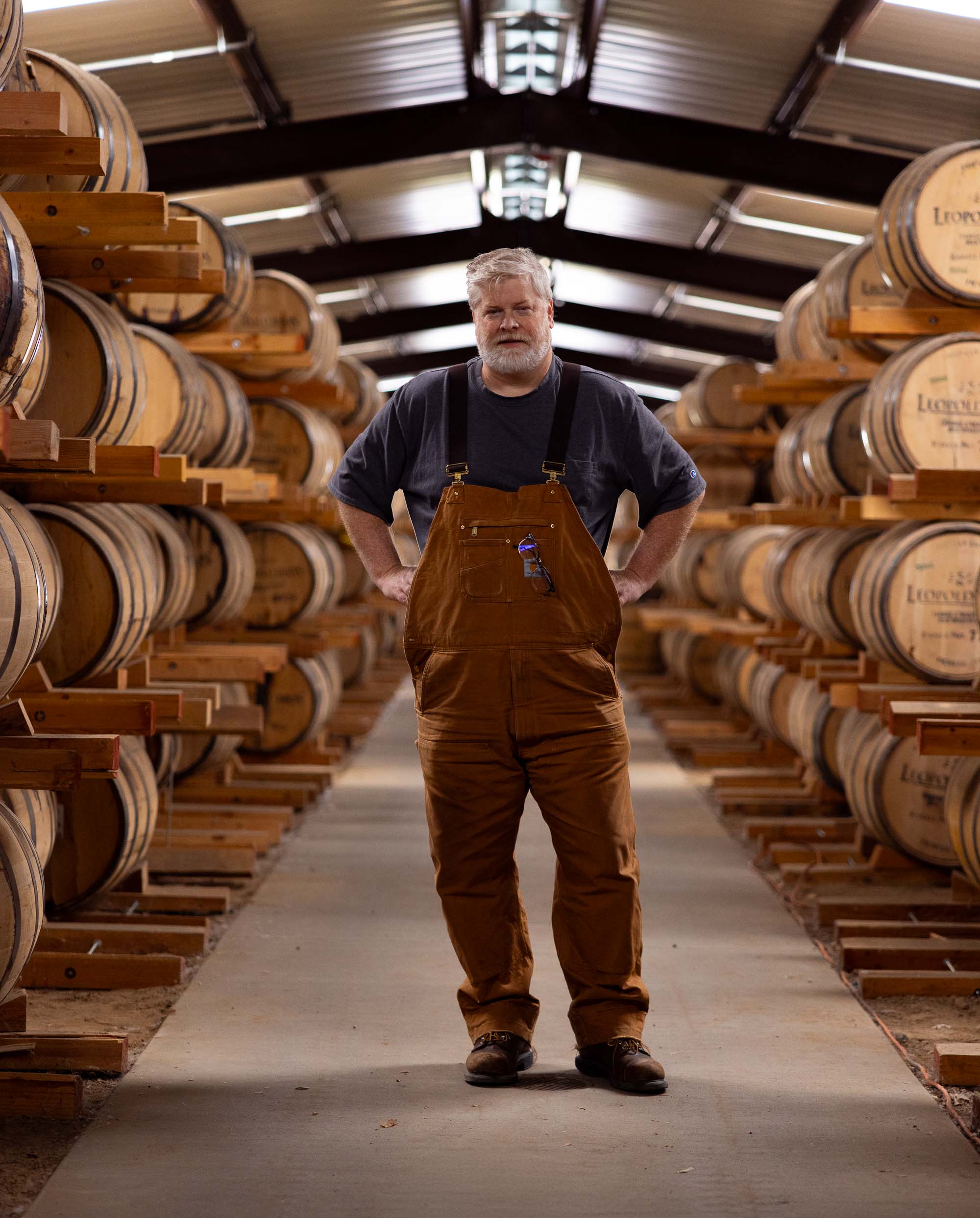 Editorial portrait of Todd Leopold, co-founder of Leopold Brothers Distillery, standing between tall stacks of whiskey barrels in the Denver aging warehouse