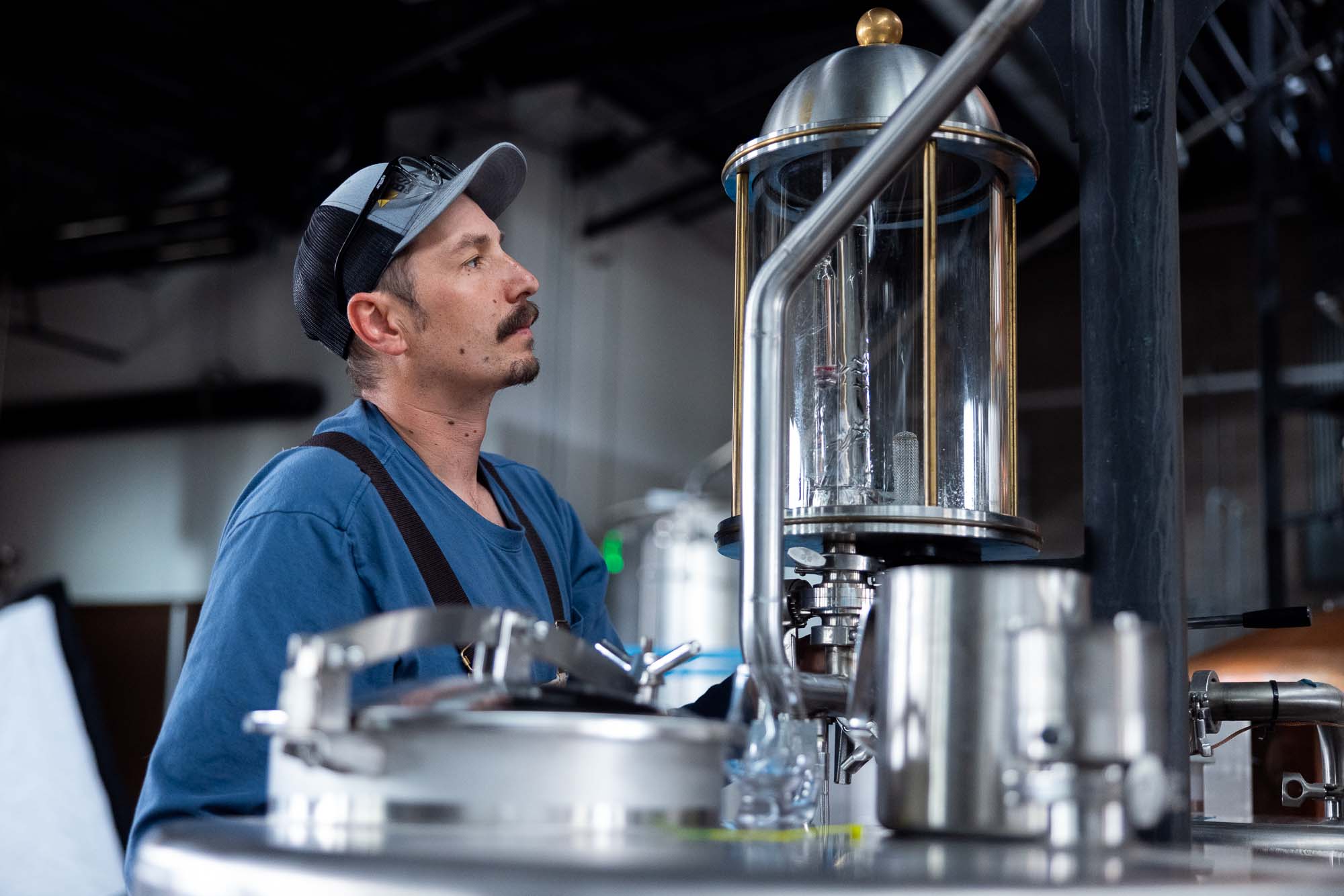 Distiller in a trucker cap inspecting the glass gin botanicals basket on top of a copper and stainless pot still during a gin run