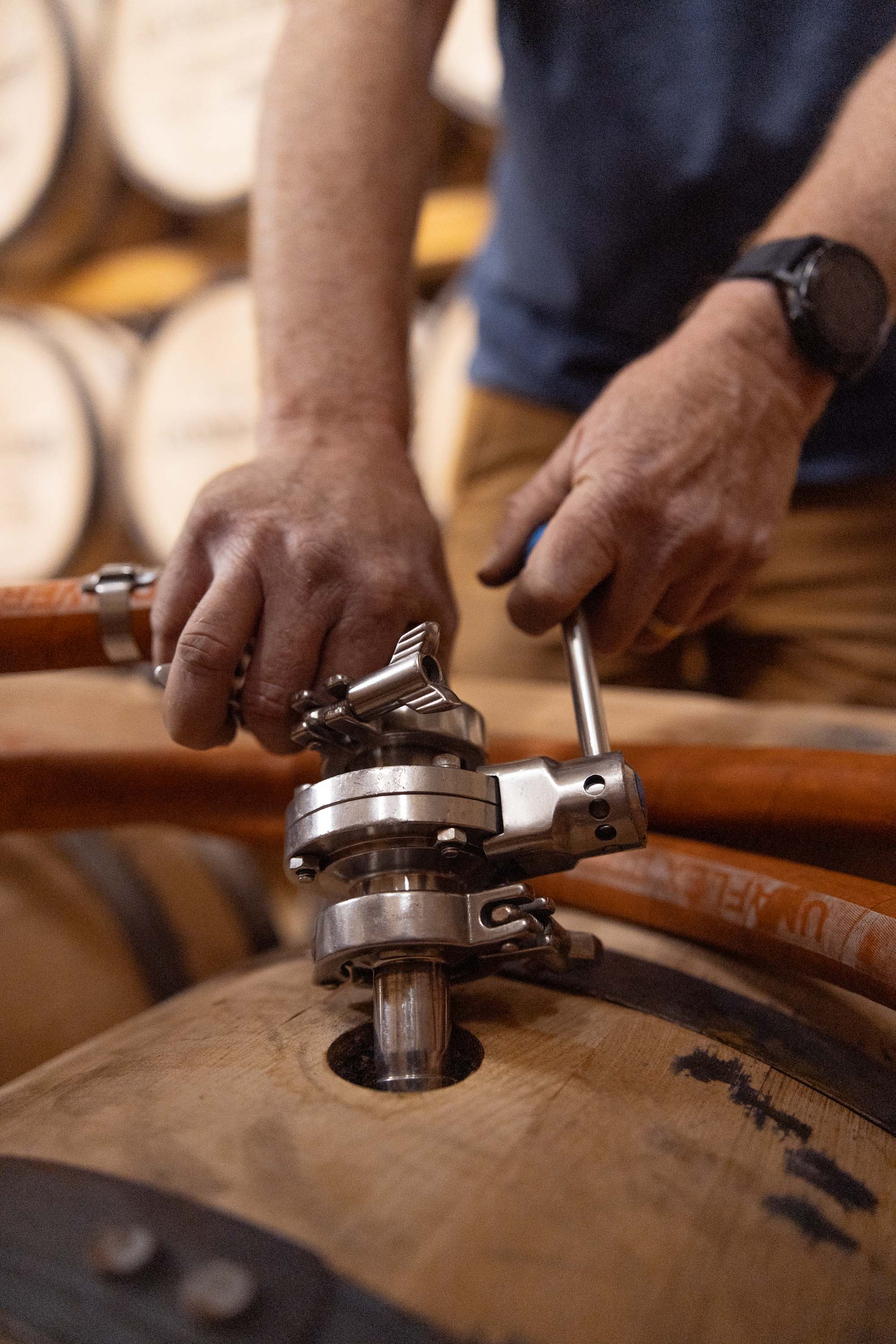 Close-up of a distiller's hands attaching a stainless steel racking valve to the bung of a Leopold Brothers whiskey barrel